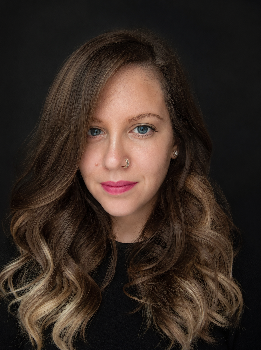 A young woman with long, wavy brown hair with blonde highlights, blue eyes, and fair skin, wearing pink lipstick, a nose ring, and small earrings, against a black background.
