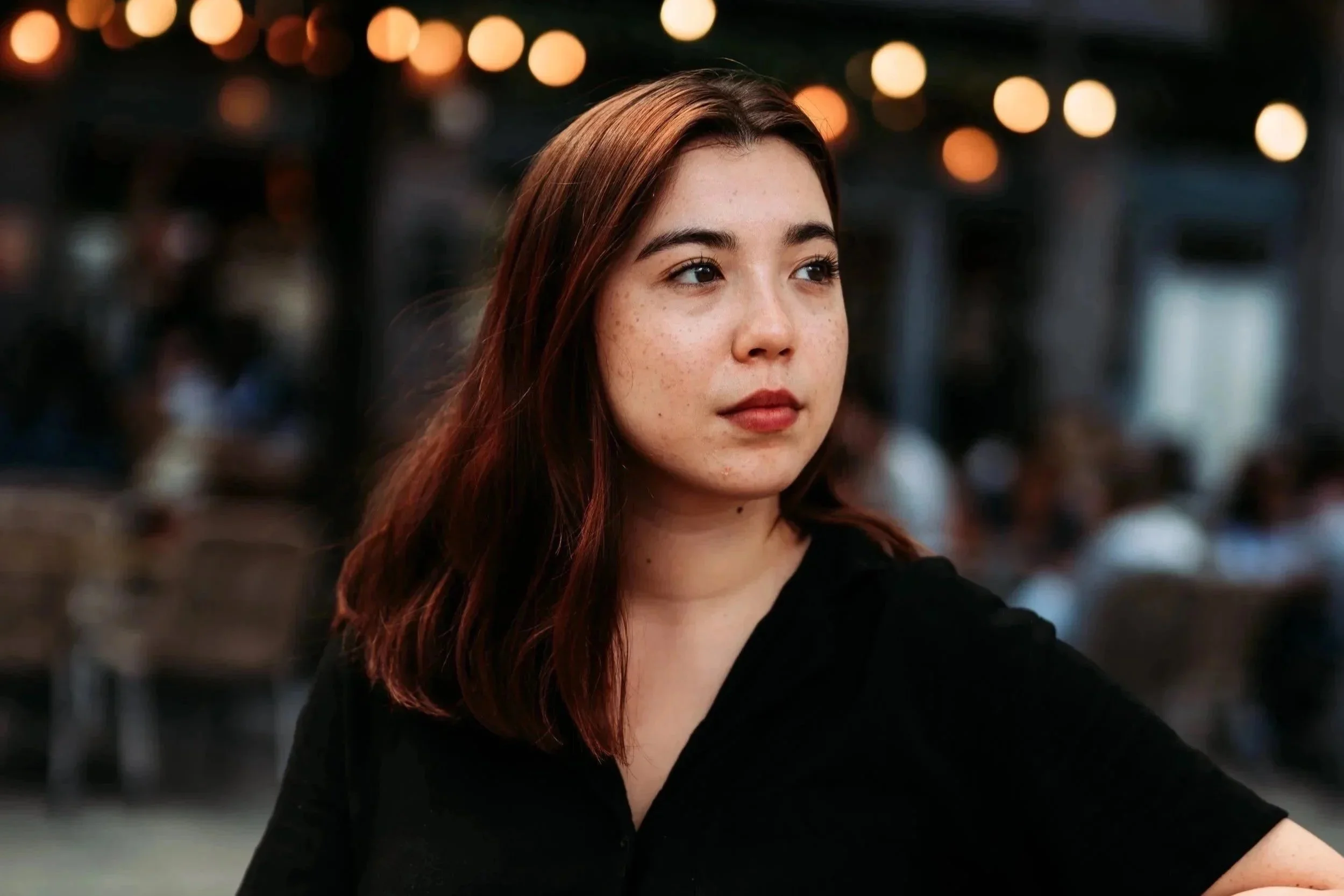 A young woman with auburn hair and freckles, wearing a black top, standing in a setting with blurred warm lights in the background.