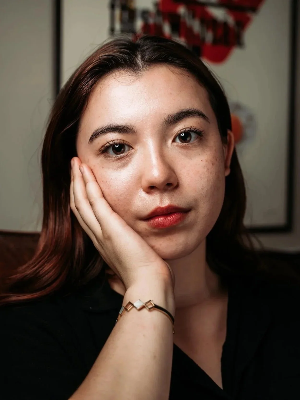 A young woman with shoulder-length dark brown hair, light skin with freckles, and hazel eyes, resting her face on her left hand, wearing a black top and a gold bracelet on her wrist.