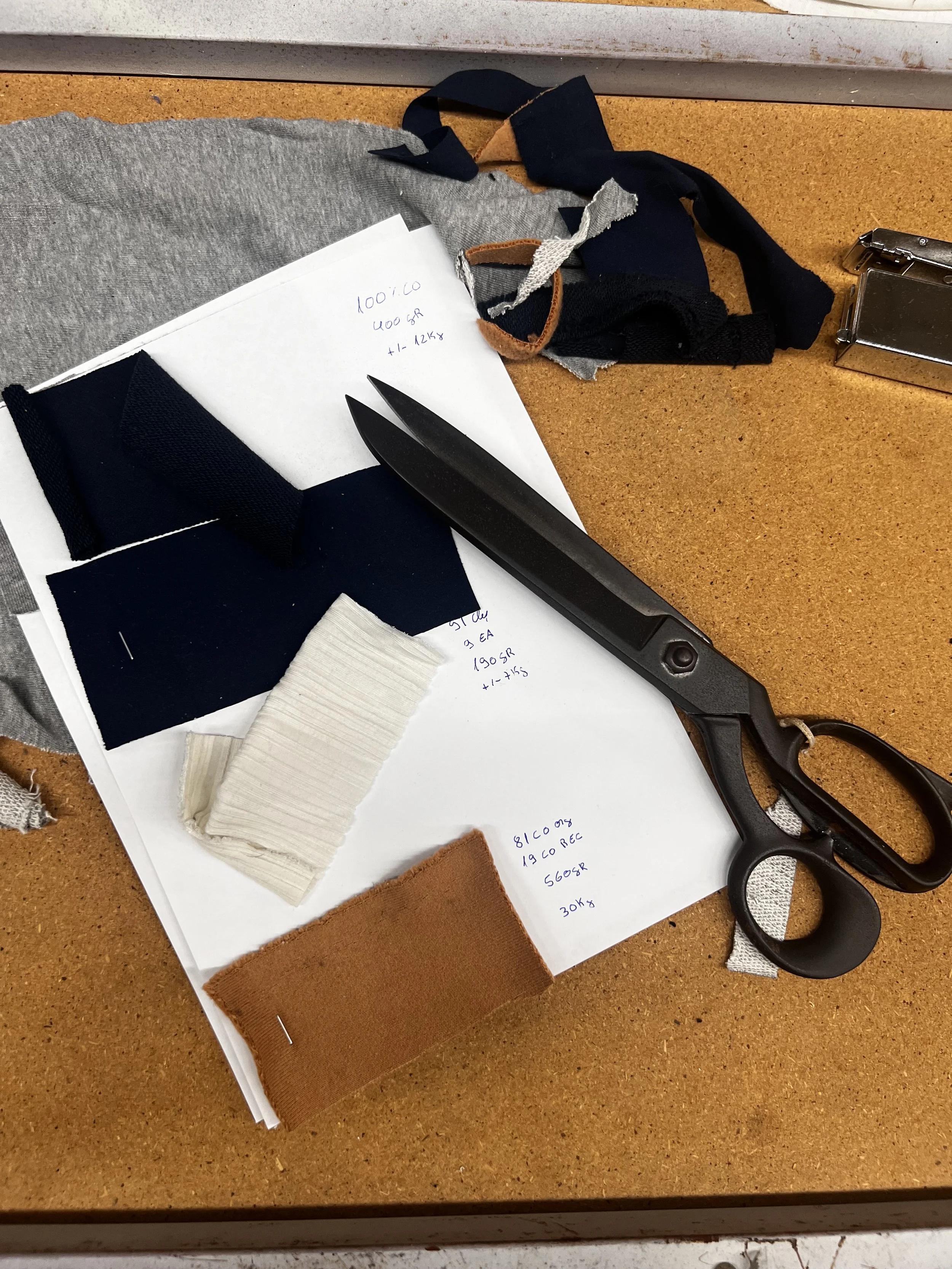 Arbeitsplatz mit Stoffproben in verschiedenen Farben und Texturen, einem schwarzen Messer, einer Schere und Notizen.