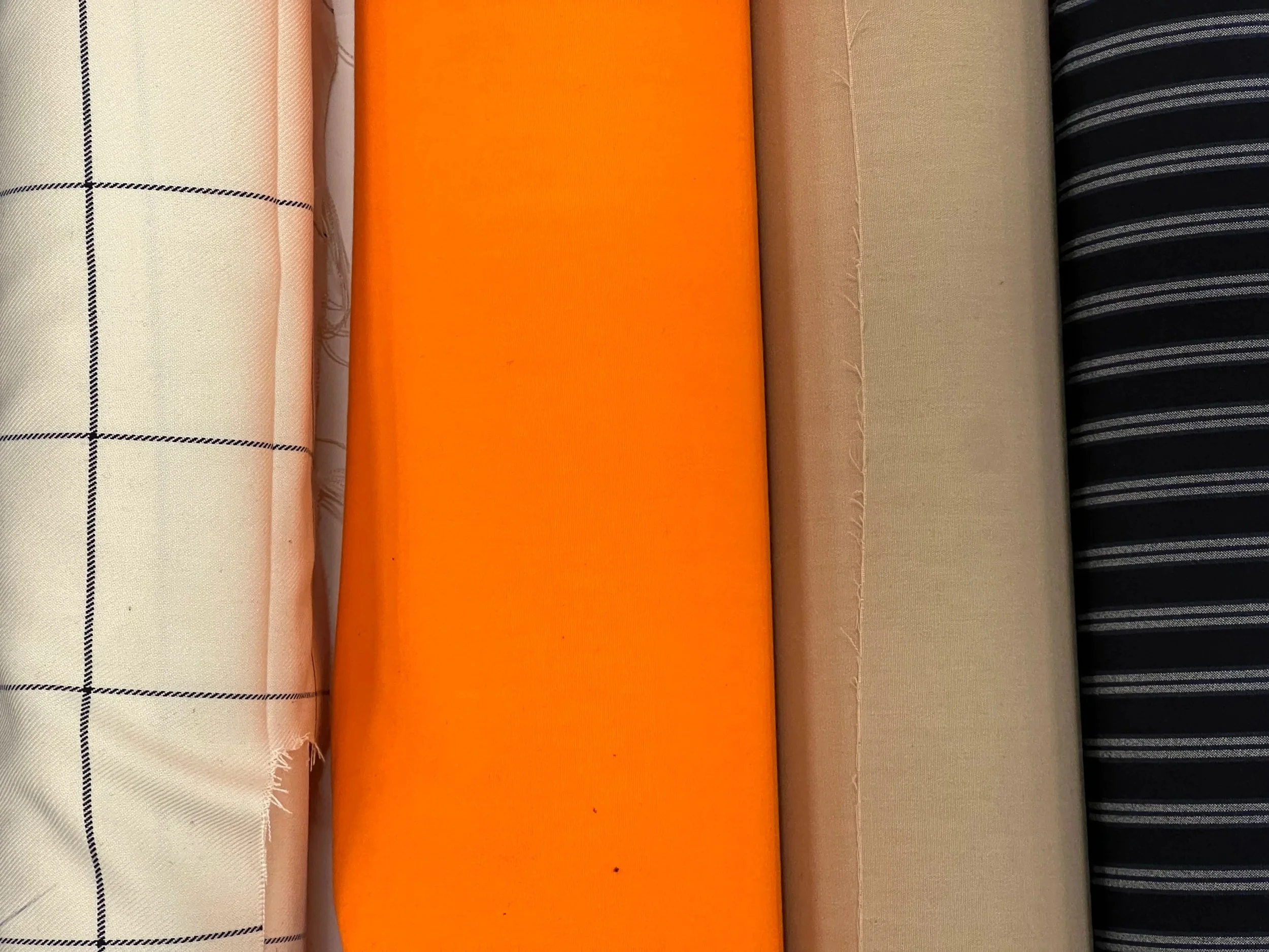 Verschiedene Rollen Stoffe mit unterschiedlichen Mustern und Farben, darunter Weiß mit Schwarzkaros, Hellfarben, Orange, Beige und Schwarz mit weißen Streifen.