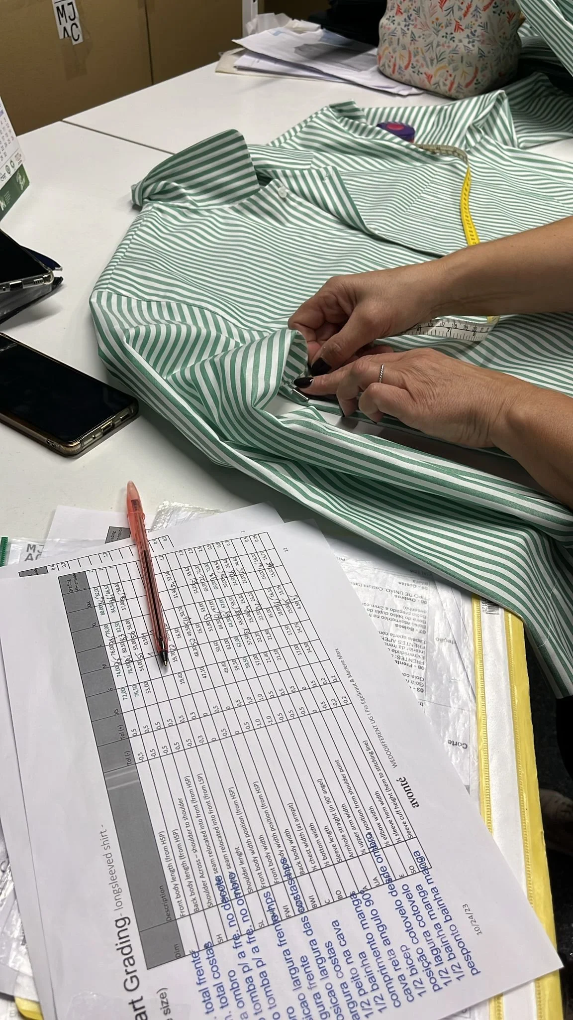 Ein Hemd mit grünen und weißen Streifen wird auf einem Tisch vermessen, mit einer Person, die es anfasst. Es liegen Papiere, ein Stift und ein Handy auf dem Tisch, im Hintergrund sind Papieren und eine Tasche sichtbar.