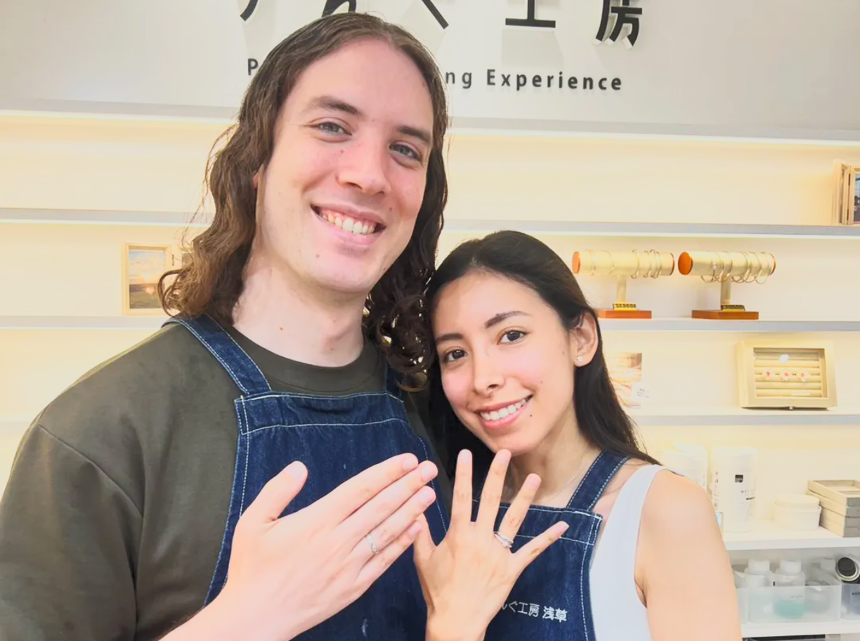 A smiling couple showing off their new rings, standing in front of a wall with Japanese characters and various small decorative items in a store.