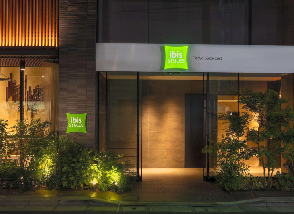 Nighttime exterior view of an Ibis Styles hotel entrance in Tokyo Ginza East, with green illuminated signs, glass sliding doors, and surrounding greenery.