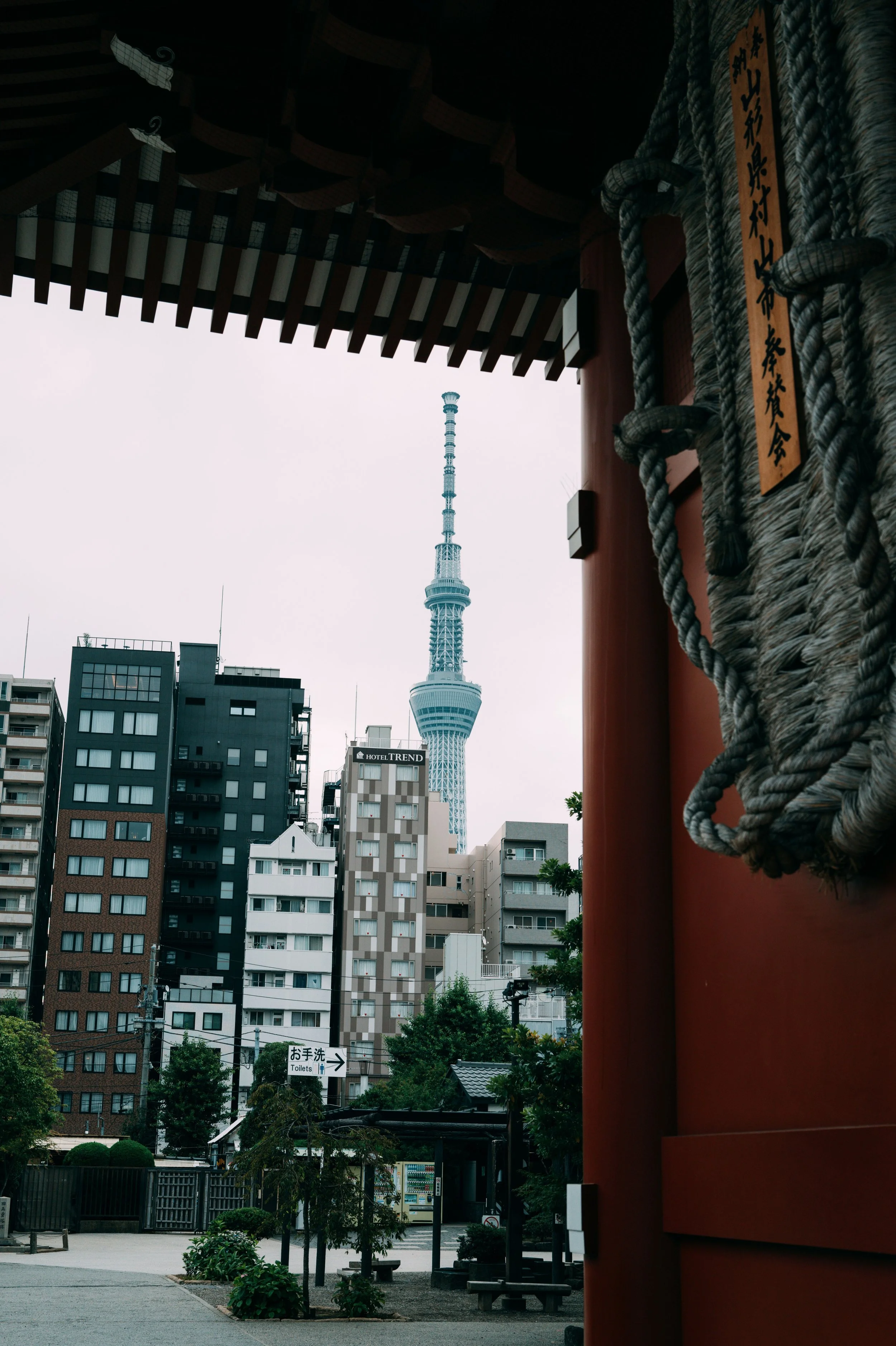 A cityscape featuring the Tokyo Tower in the distance, with modern buildings in the foreground. The image is taken from a traditional Japanese structure, with part of a red-orange wall and thick ropes visible on the right side.