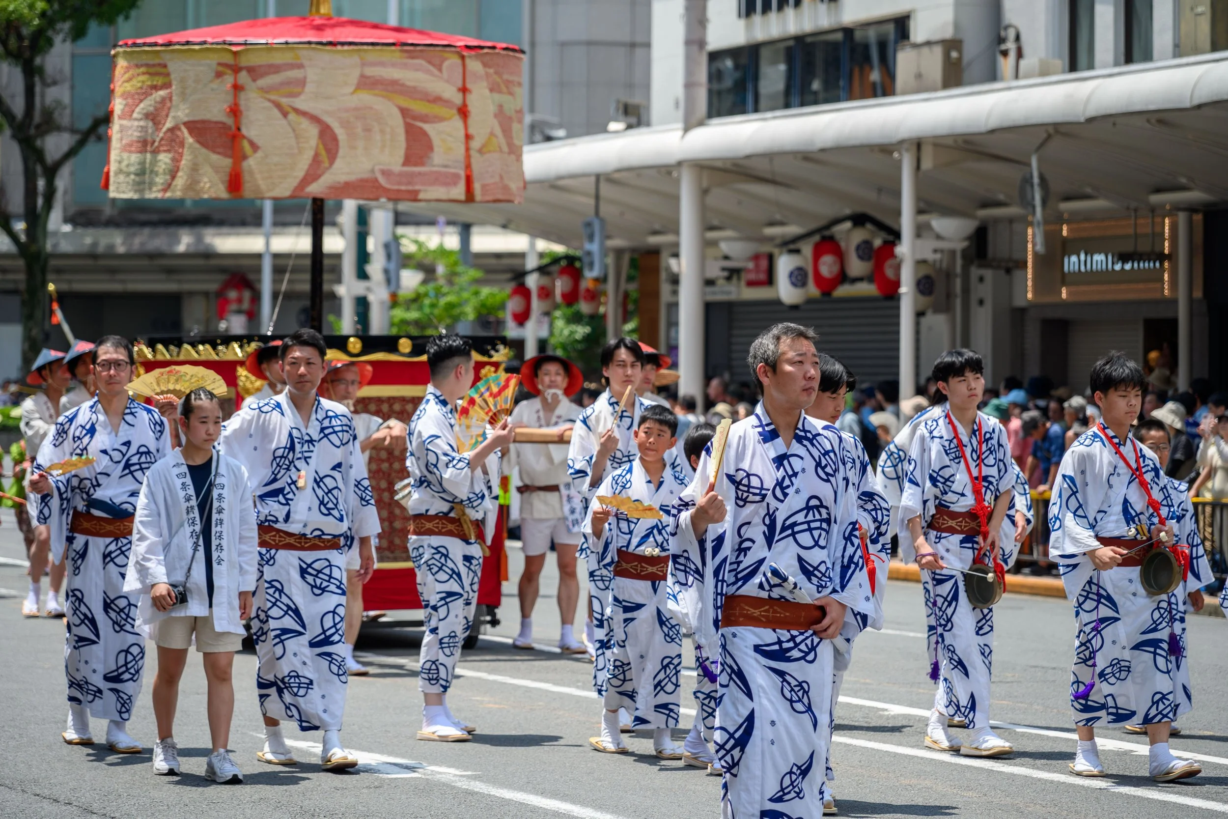 A group of people participating in a traditional Japanese festival parade, wearing matching white and blue patterned yukata and carrying fans, with a float decorated with lanterns in the background.