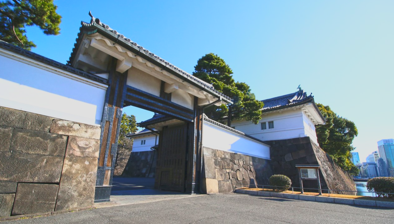 Traditional Japanese castle gate with white walls, black tiled roof, and stone foundation, surrounded by greenery under a clear blue sky.