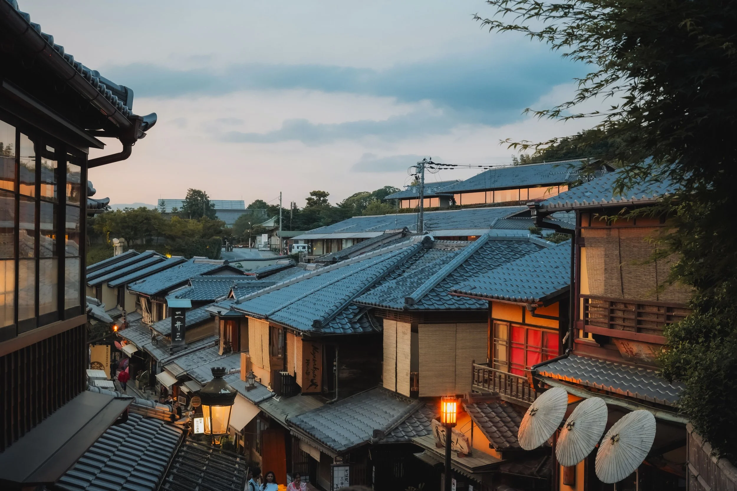 Traditional Japanese buildings with tiled roofs at dusk, some illuminated by warm lights, and people walking through a narrow street.