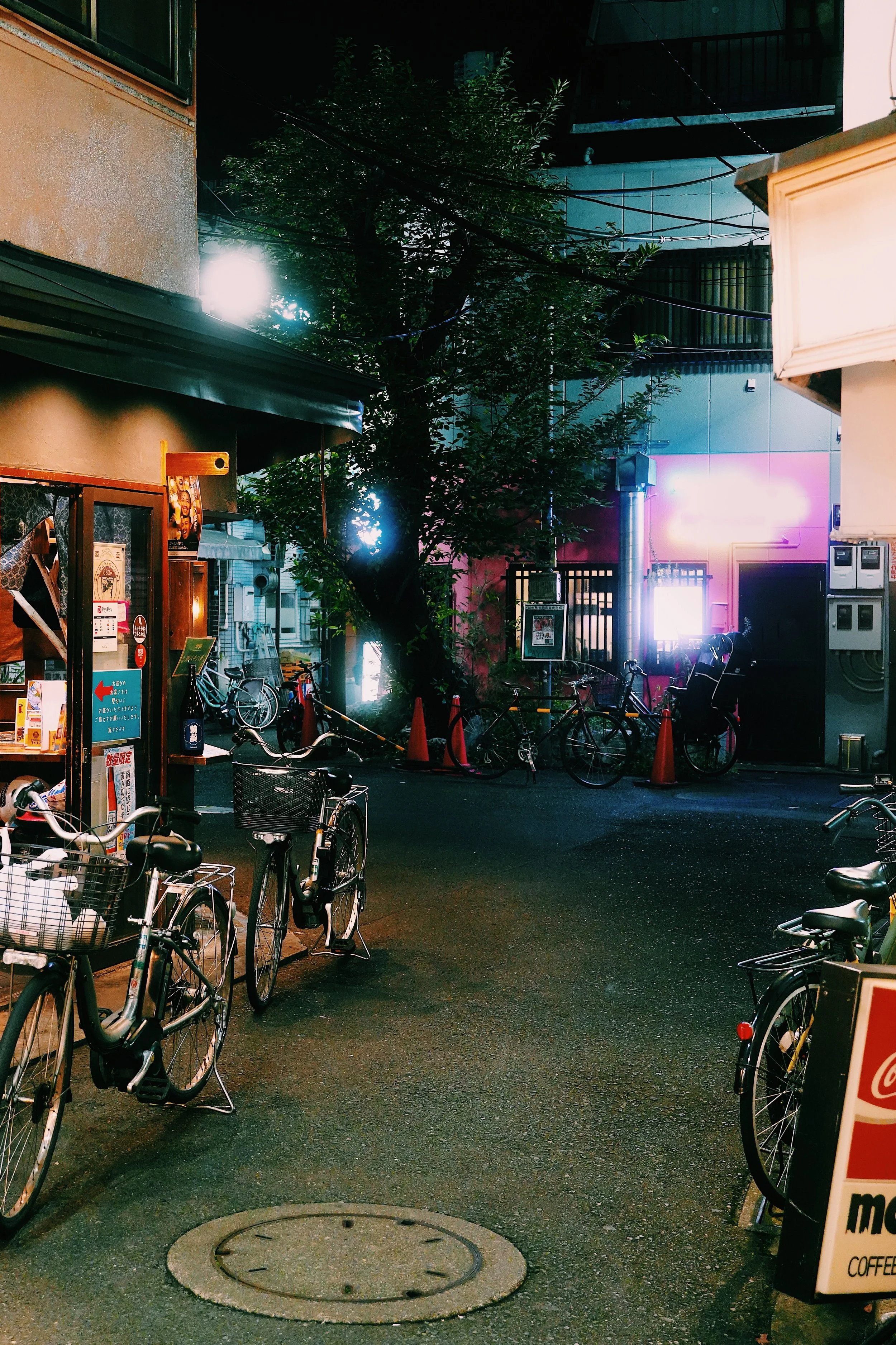 Night scene of a street with parked bicycles, a tree, and illuminated signs. Bicycles are aligned along the sidewalk near a building with various posters and signs. Bright lights and neon signs are visible in the background, creating a vibrant urban atmosphere.