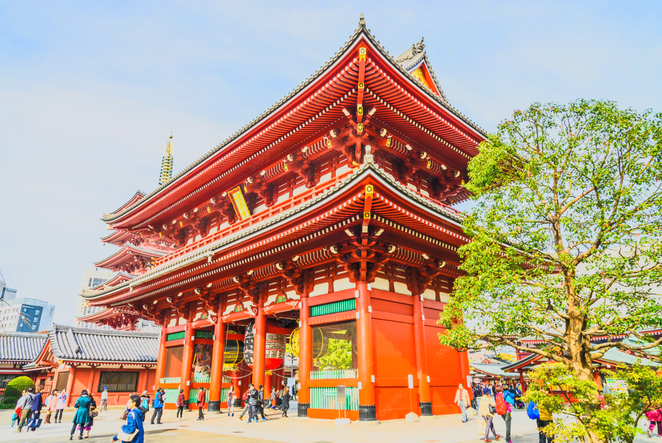Traditional Japanese temple with red wooden structure, ornate roofing, and a surrounding courtyard with visitors, trees, and city buildings in the background.