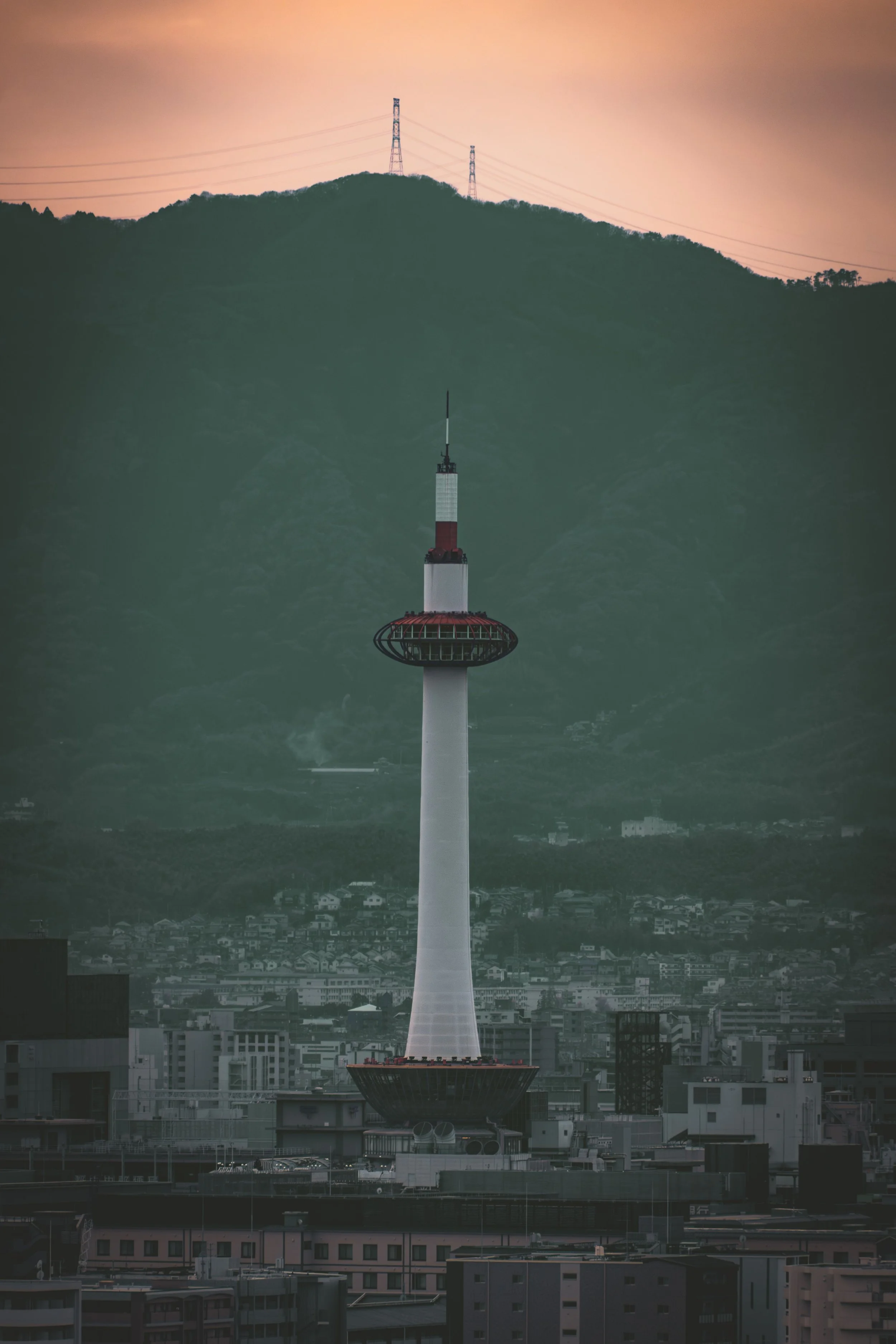 A cityscape at dusk featuring the Tokyo Skytree, a tall slim tower with a red and white structure, set against a backdrop of a mountainous terrain with power lines running across the hills.
