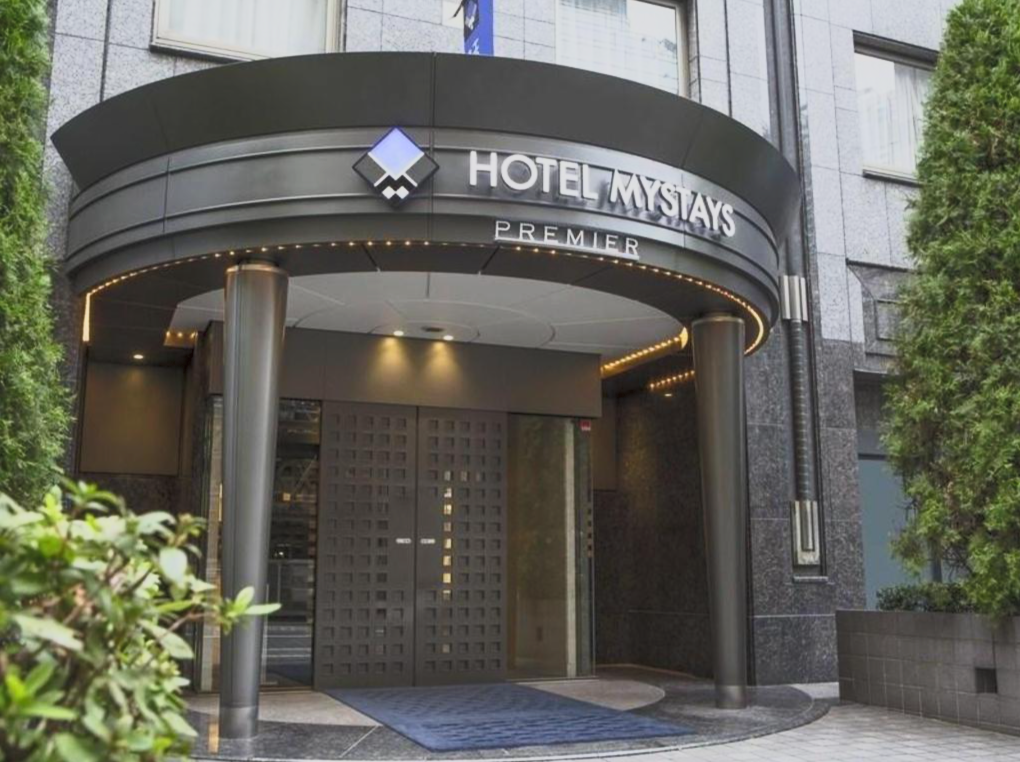Entrance of Hotel Mystays Premier with a black archway, illuminated lights, and a patterned double door, surrounded by greenery.