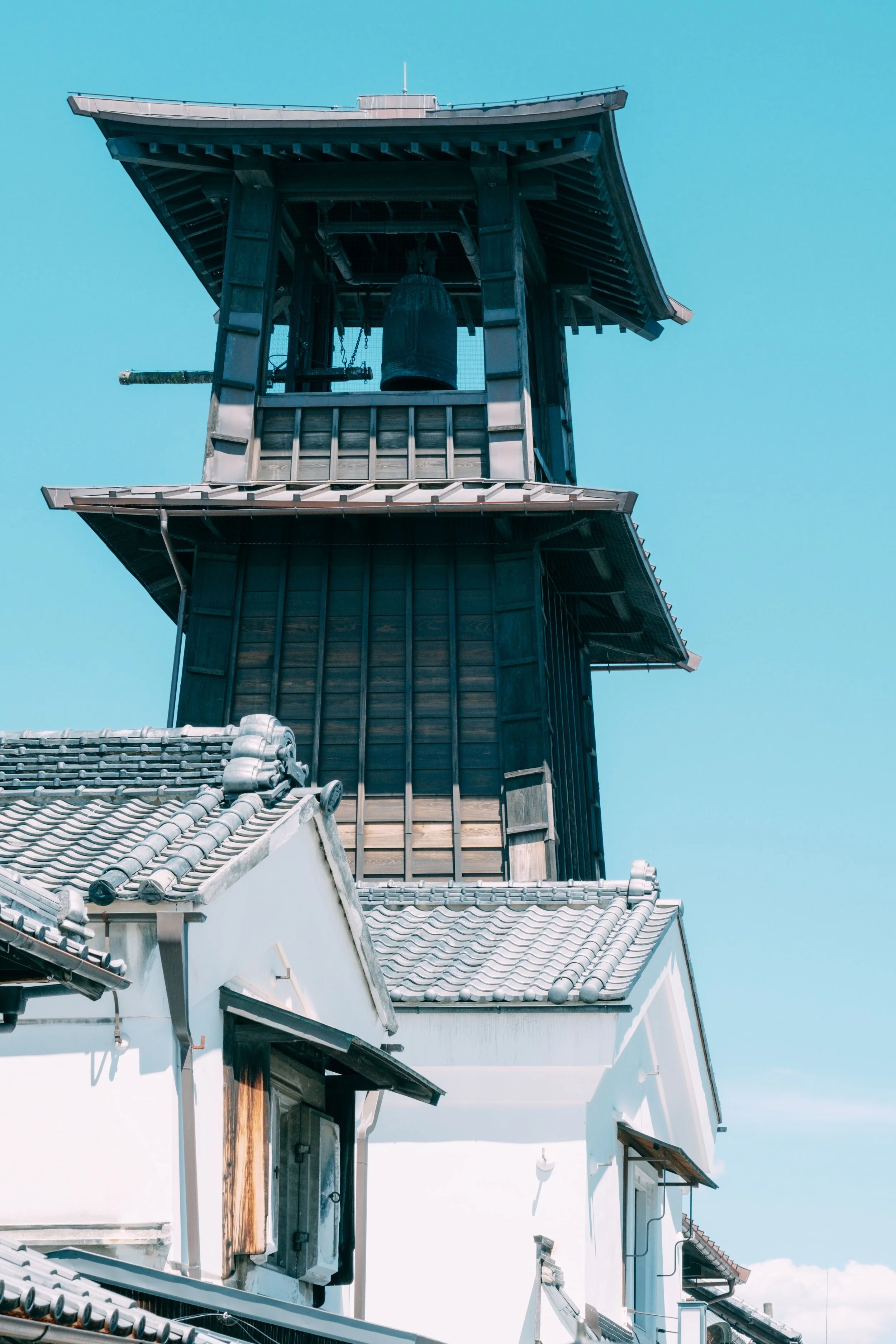 A traditional Japanese building with a wooden bell tower, tiled roof, and white walls against a blue sky.