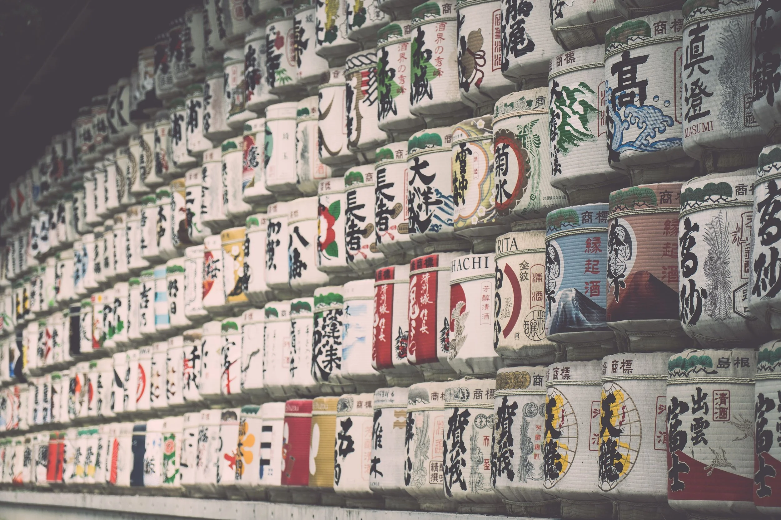 Rows of Japanese sake barrels with colorful, traditional calligraphy and illustrations stacked on shelves.