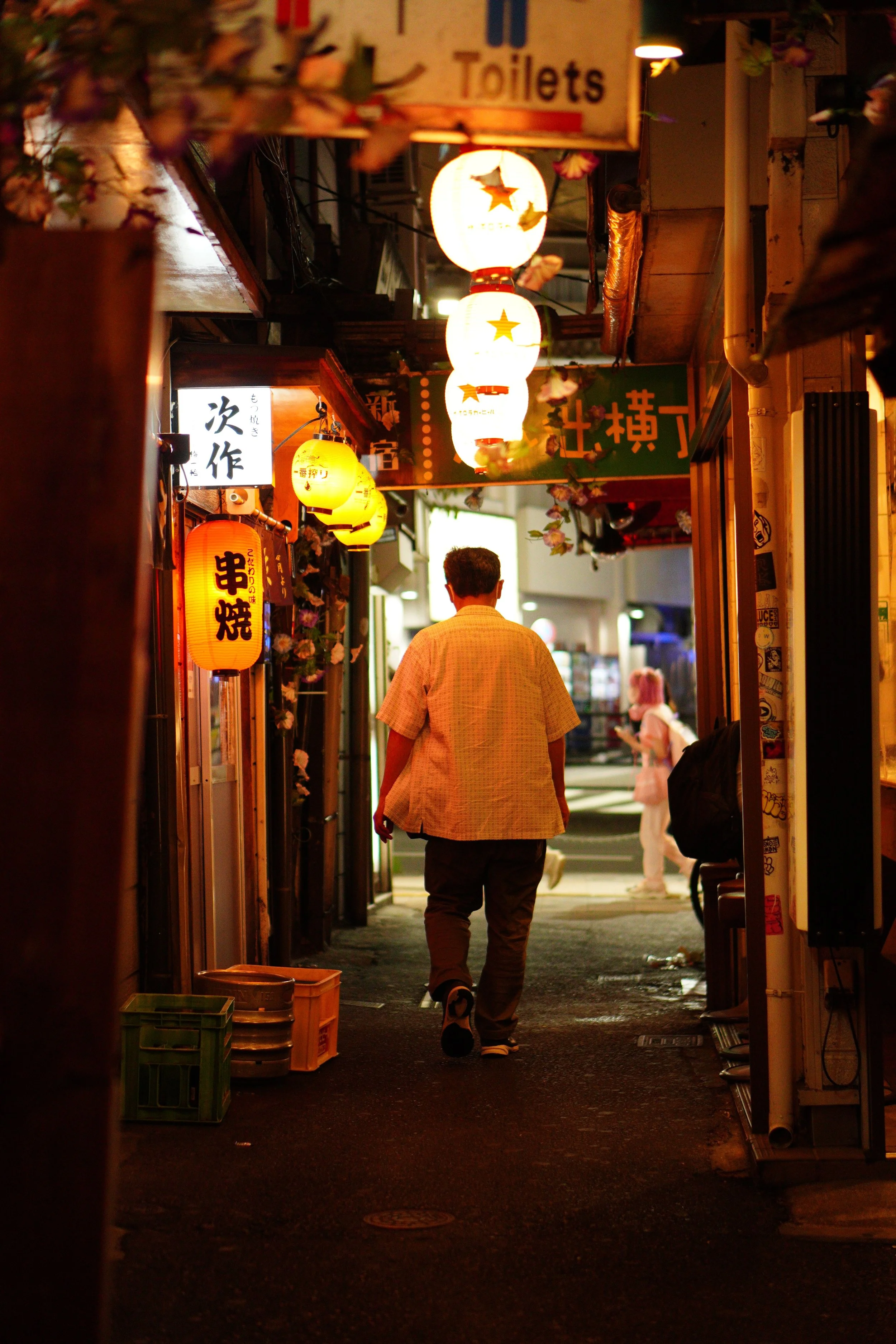 Night scene in a narrow street illuminated by hanging paper lanterns and signs, with a man walking away from the camera and a woman in the background.