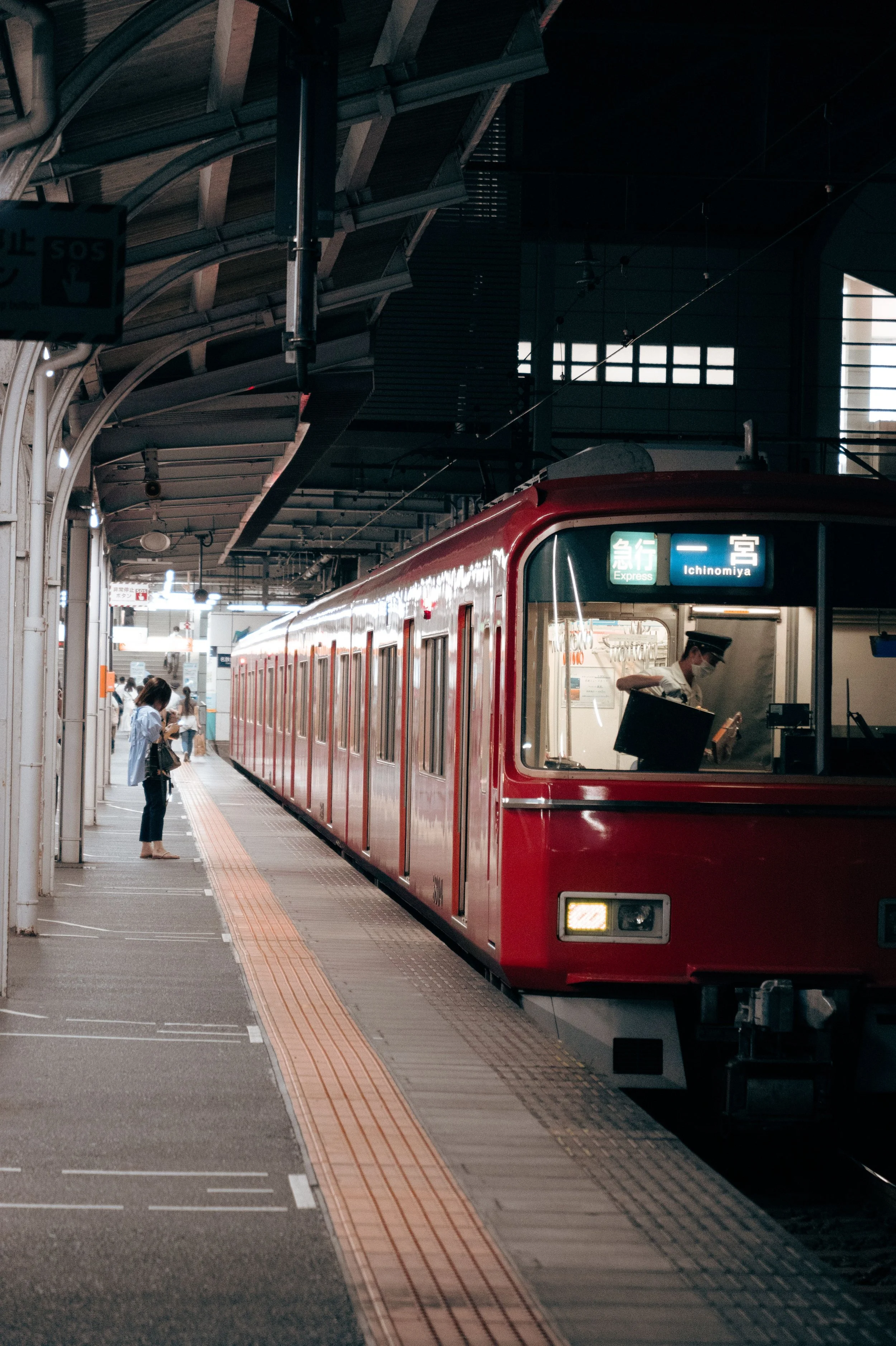 A red train at a station platform with passengers, some wearing masks, waiting, and a sign indicating the train to Ichinomiya.