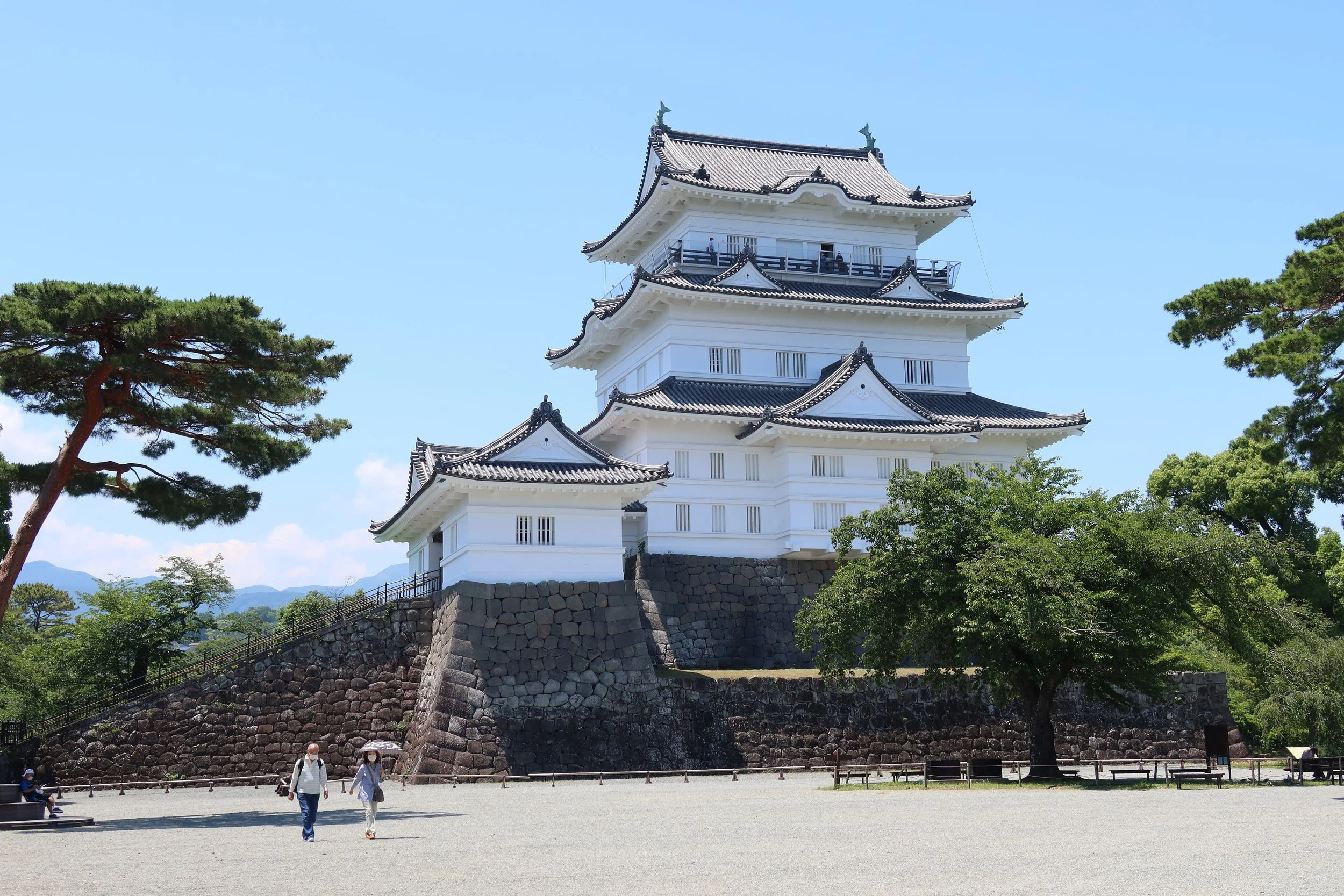 Traditional Japanese castle with white walls and black tiled roofs, surrounded by green trees and a stone wall base, with visitors walking nearby under a clear blue sky.