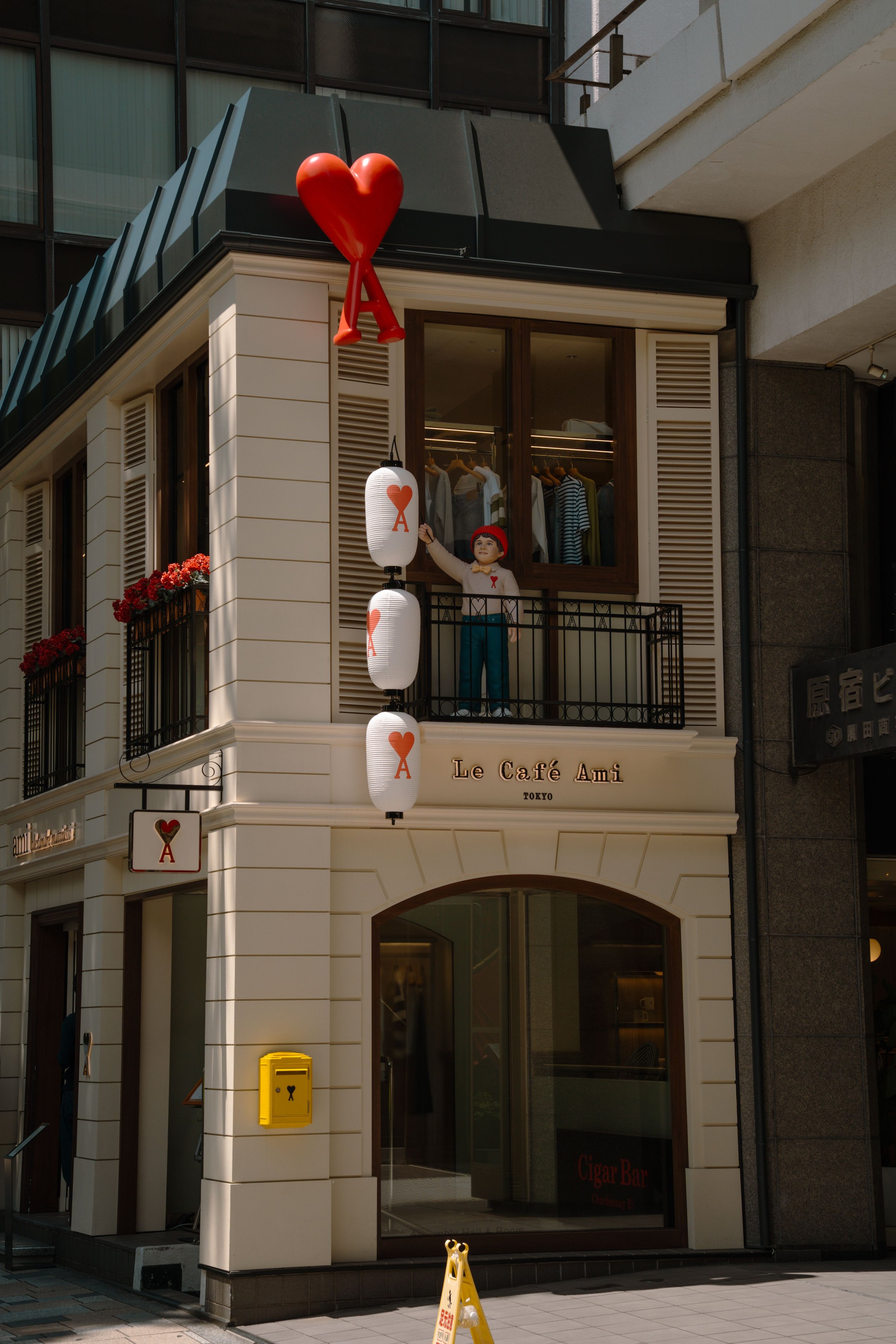 Building with white exterior, black railing, red hearts, paper lanterns with hearts, and signage reading "Le Café Ami" and "Cigar Bar" in Tokyo.