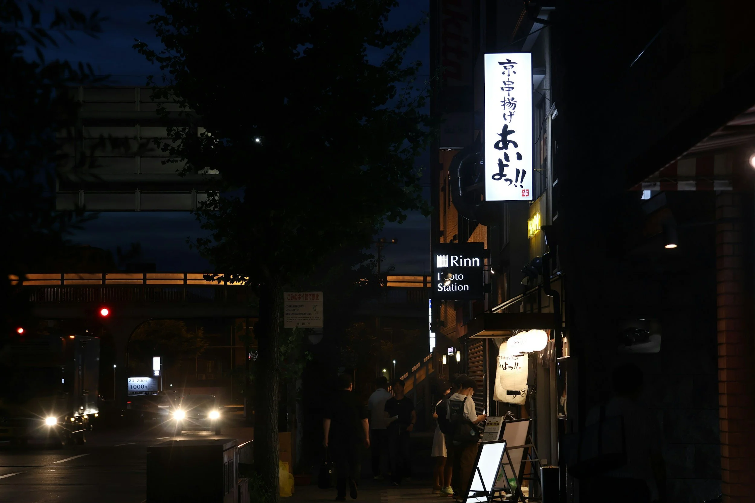 Nighttime street scene with illuminated signs in Japanese, pedestrians, and vehicles with headlights on.