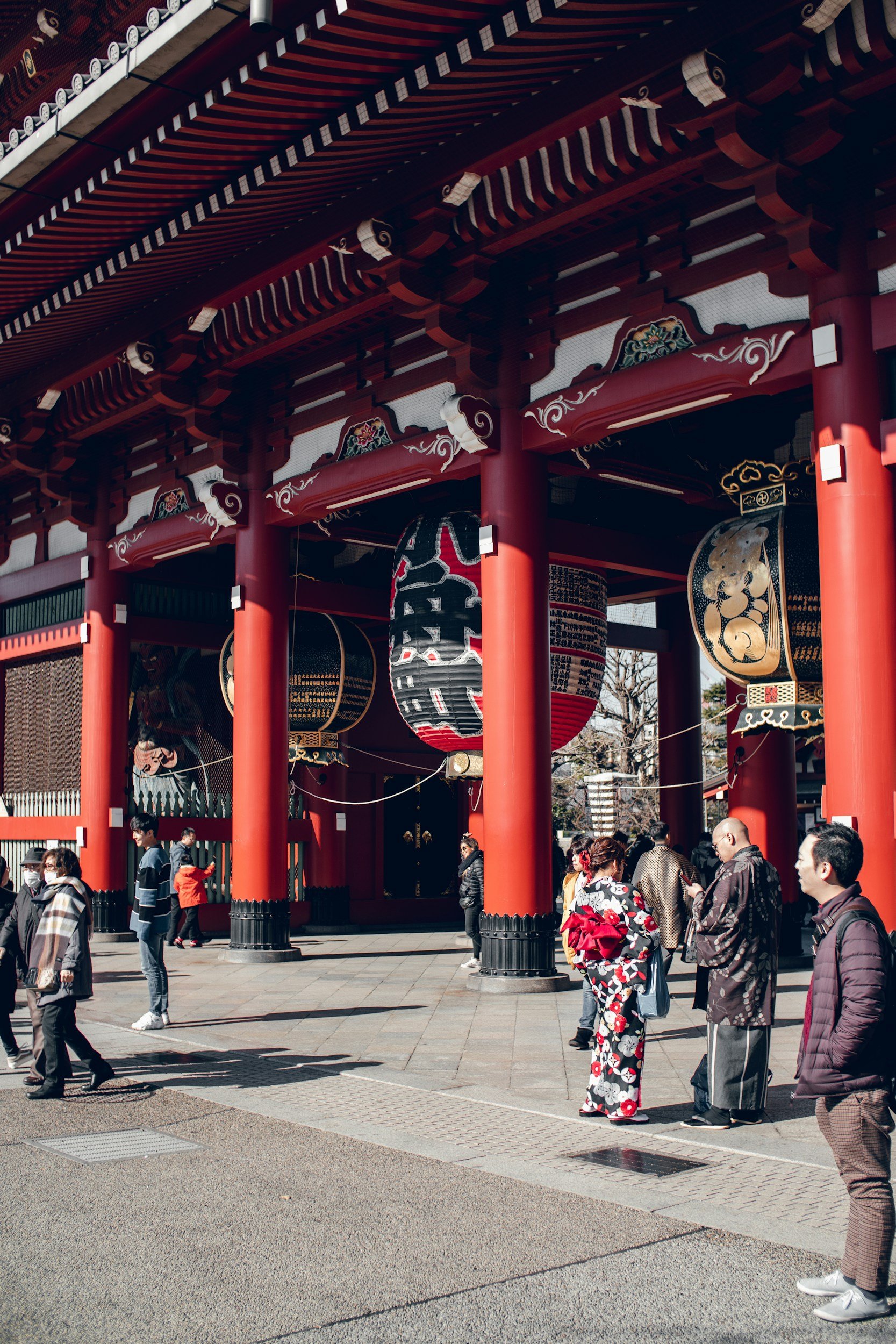 People in traditional Japanese kimonos and modern attire gather in front of a large, ornate red temple gate with black and gold accents, decorative lanterns, and intricate architectural details.