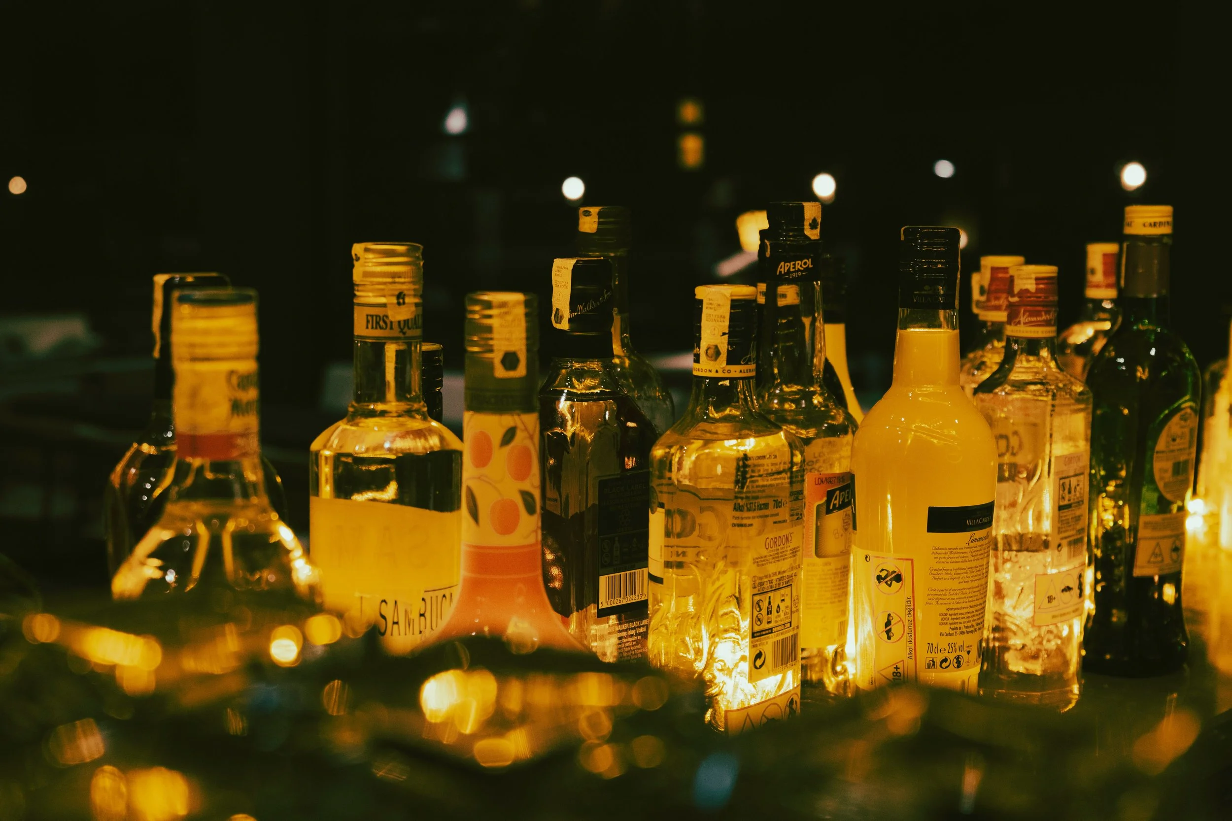 A bar shelf illuminated with warm yellow lights, displaying various bottles of liquor, including whiskey, vodka, and liqueurs, with some labels visible.