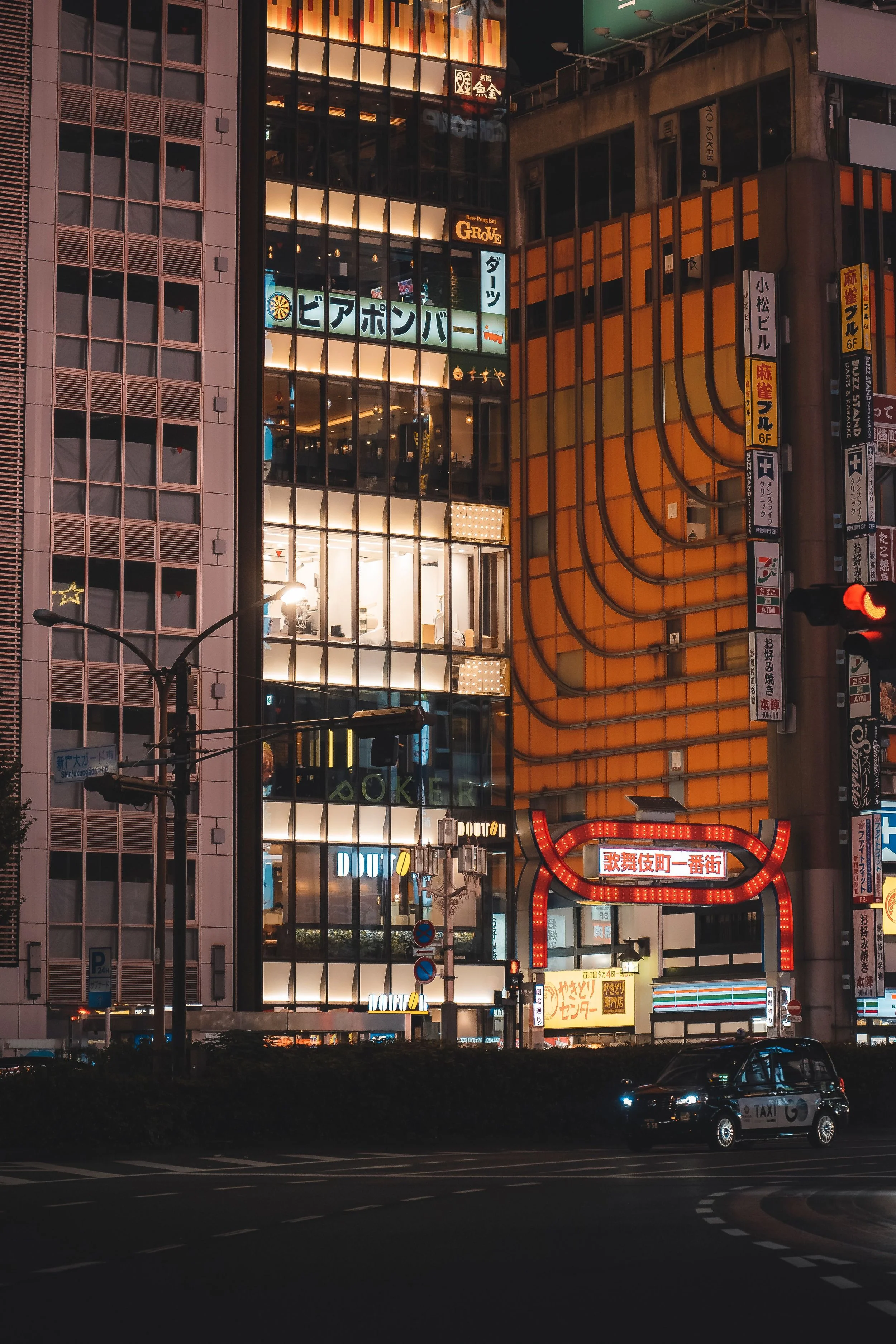 View of a city street at night with illuminated signs in Japanese, including a rounded red lighted sign at the bottom and a taxi driving on the road in the foreground.
