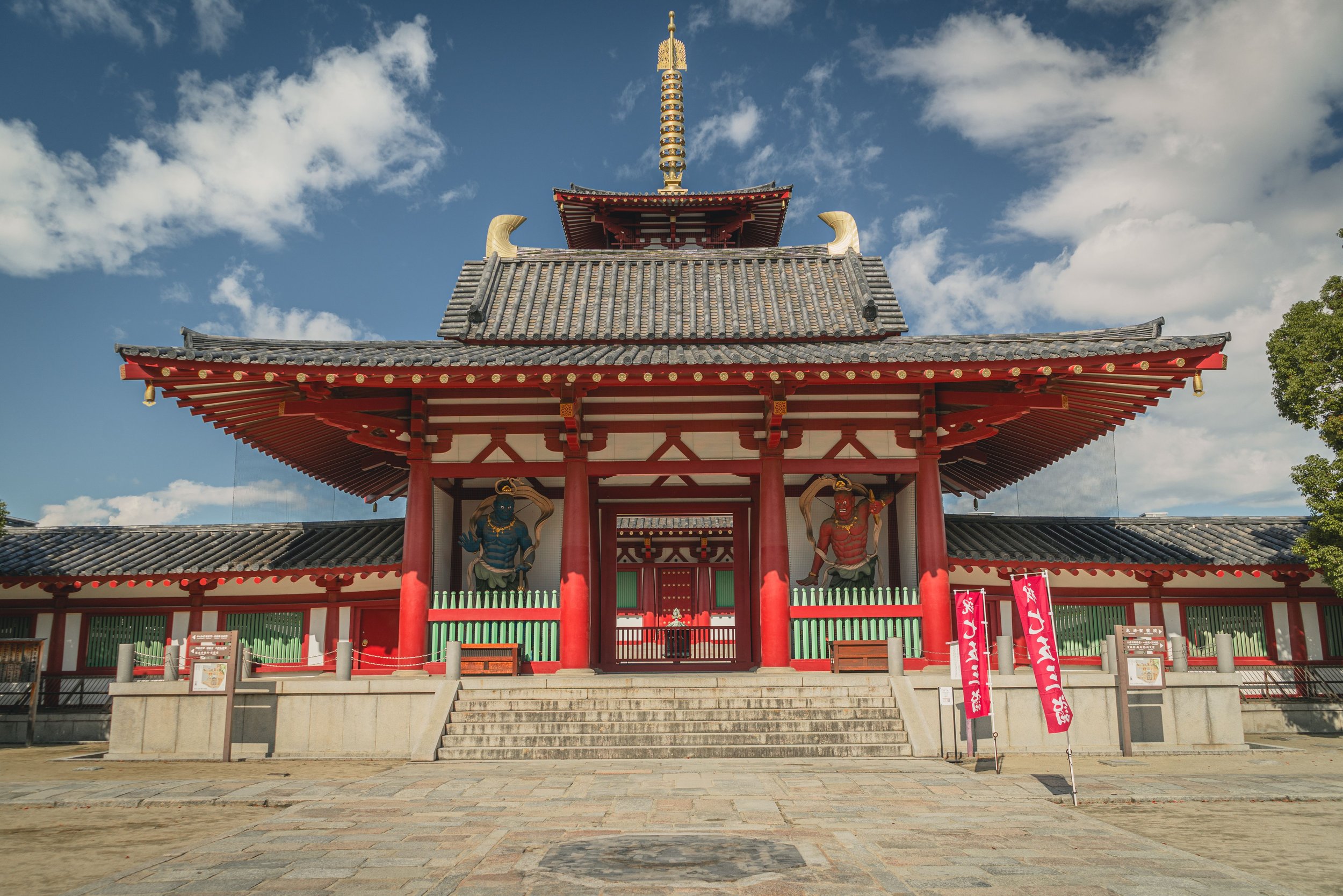 Traditional Japanese temple structure with a tiered roof, red and white walls, steps leading up to it, and statues of mythological creatures at the entrance. The sky is partly cloudy with blue skies and white clouds.