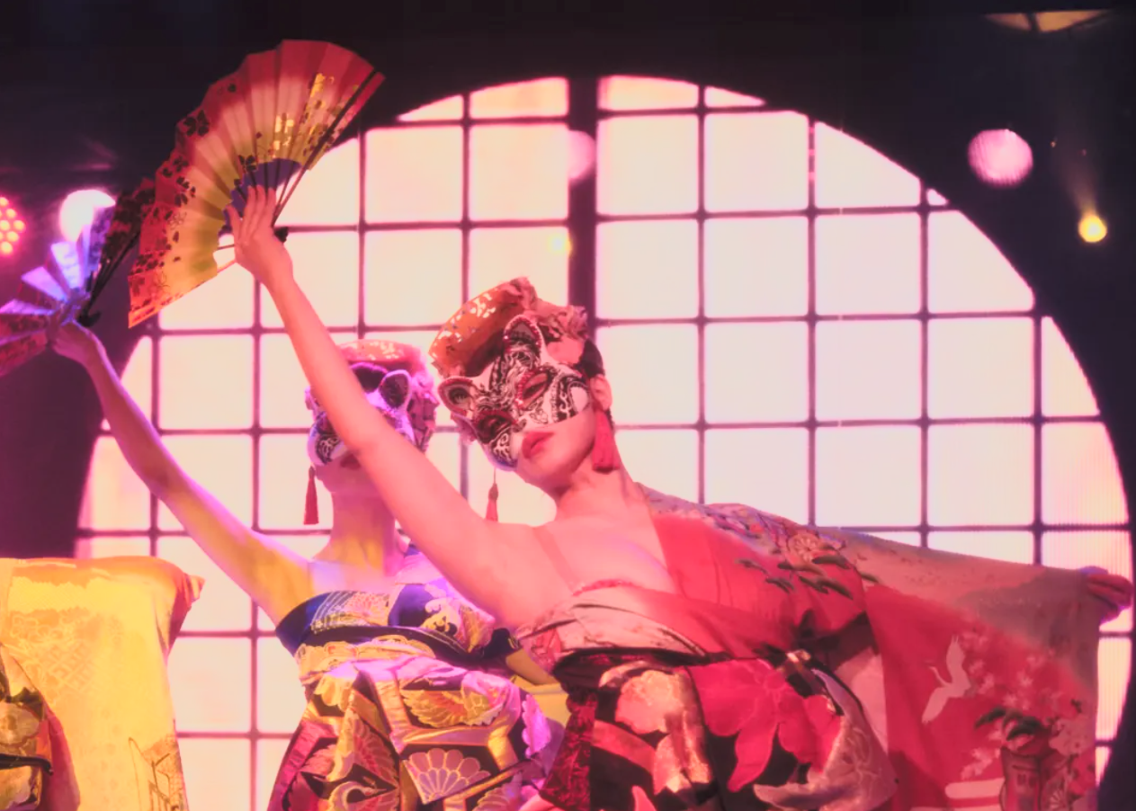 Two performers wearing ornate masks and colorful kimonos, holding fans, performing in front of a large illuminated window.