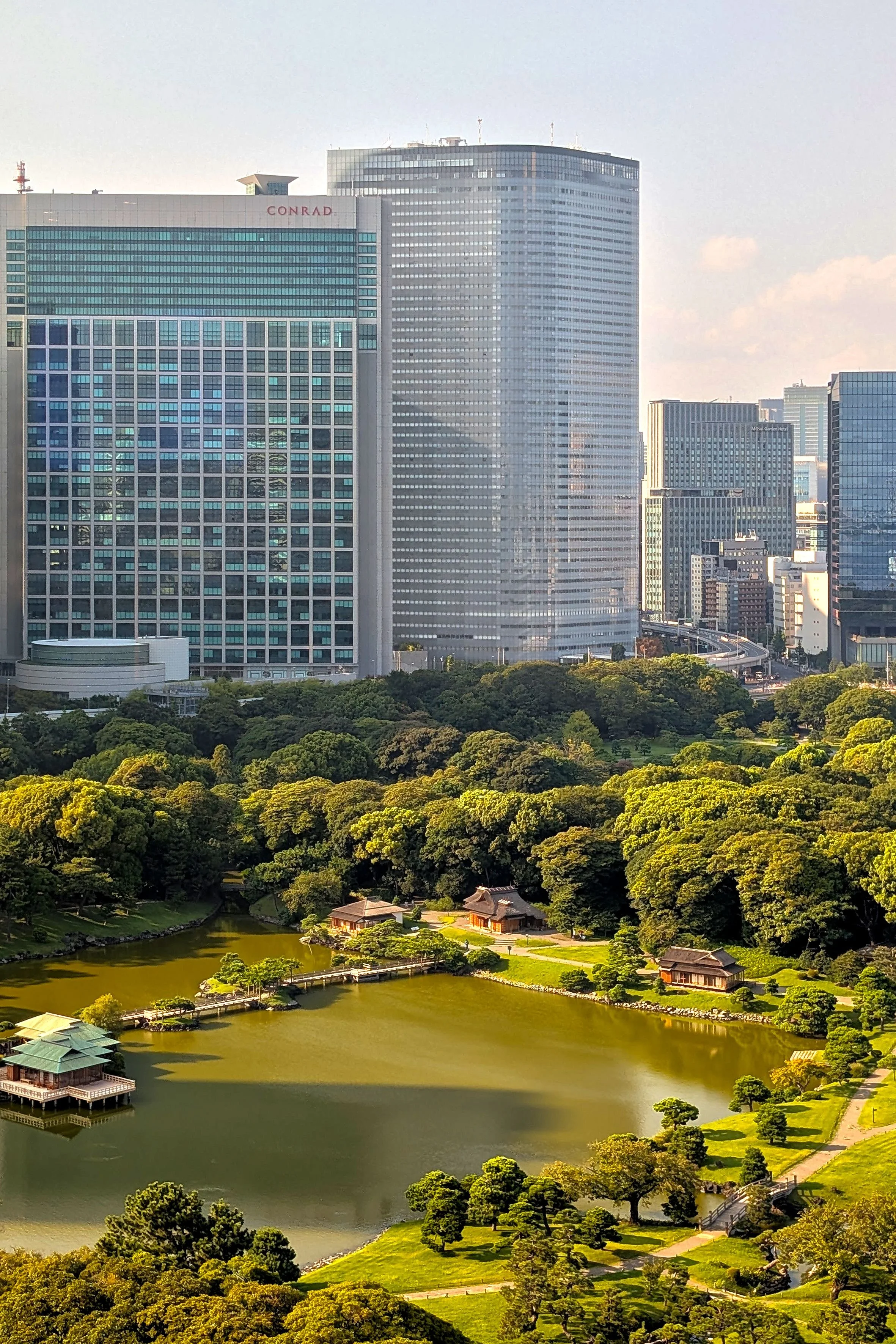 Panoramic view of a city skyline with tall modern buildings in the background and a lush traditional Japanese garden with a pond, trees, and small traditional buildings in the foreground.