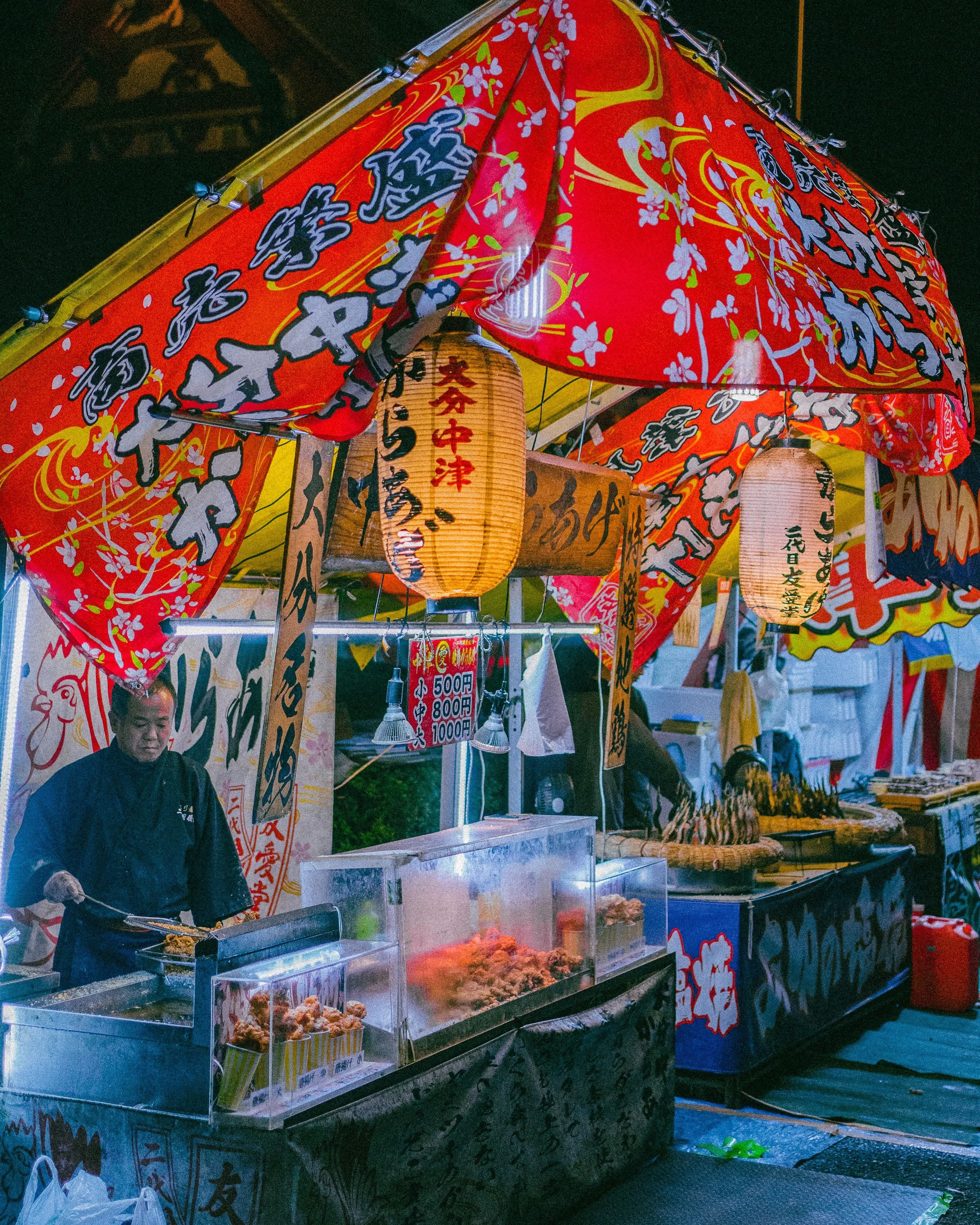 A food stall at night in Japan selling fried chicken, with a man in traditional attire preparing food. The stall is decorated with red, yellow, and white banners, paper lanterns, and signs displaying prices.