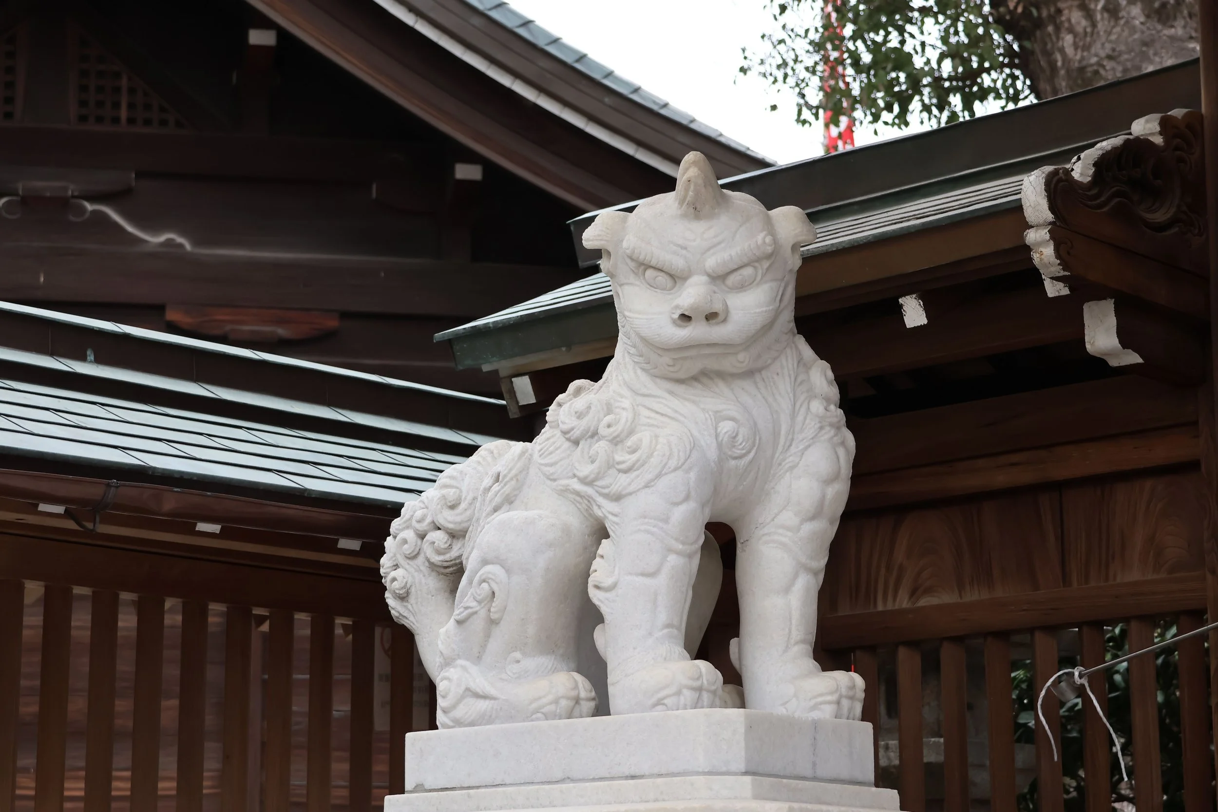 White stone statue of a mythical creature, possibly a guardian lion or foo dog, on a pedestal outside a wooden structure with a metal roof and a tree in the background.