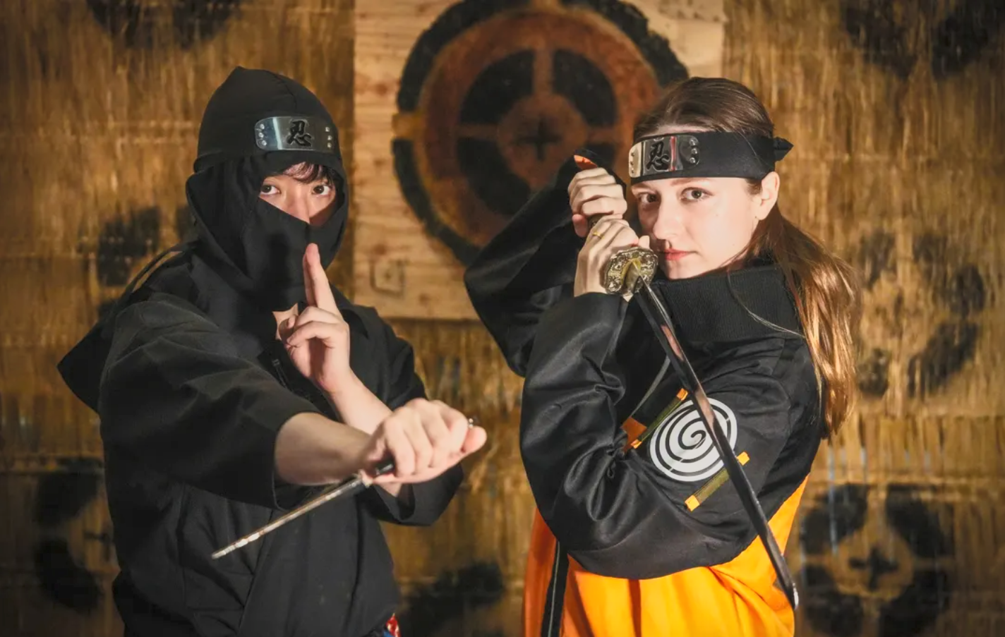 Two women posing as ninjas, one in black with a covered face and the other in black and orange, holding katanas in a dojo with wooden walls and target in the background.