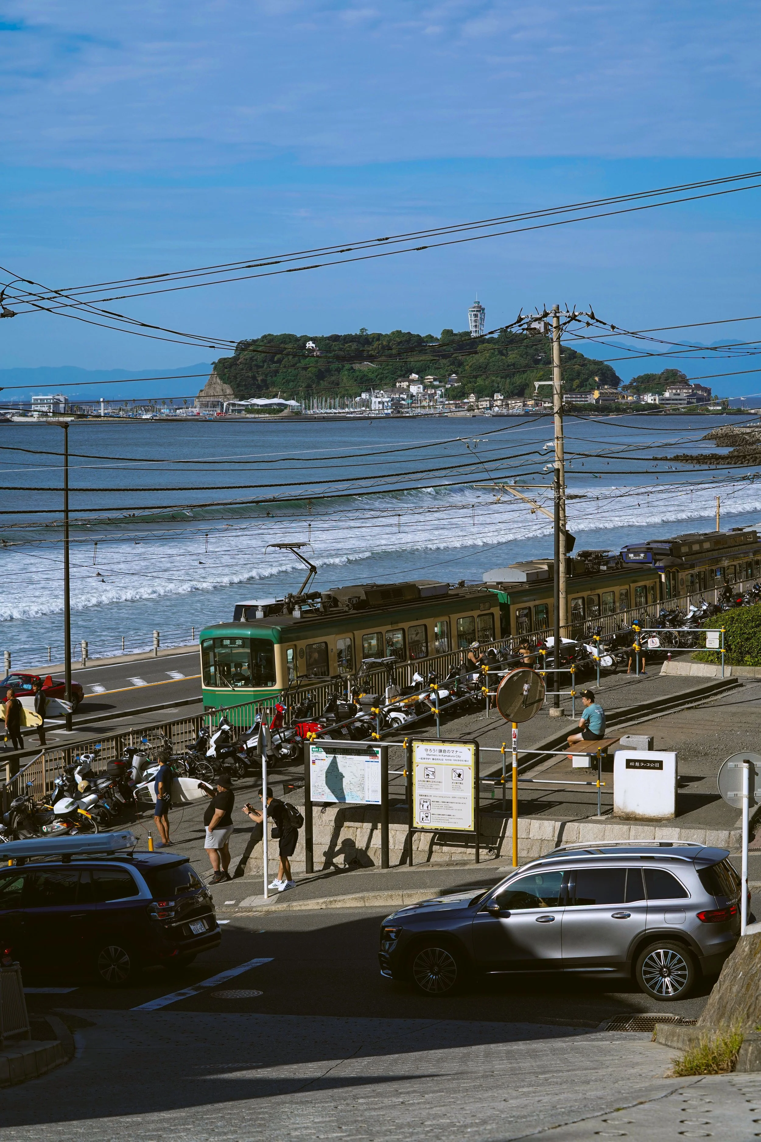 People waiting and walking near a street with cars, a tram, and electrical poles; in the background, a coastal hill with a lighthouse and buildings.