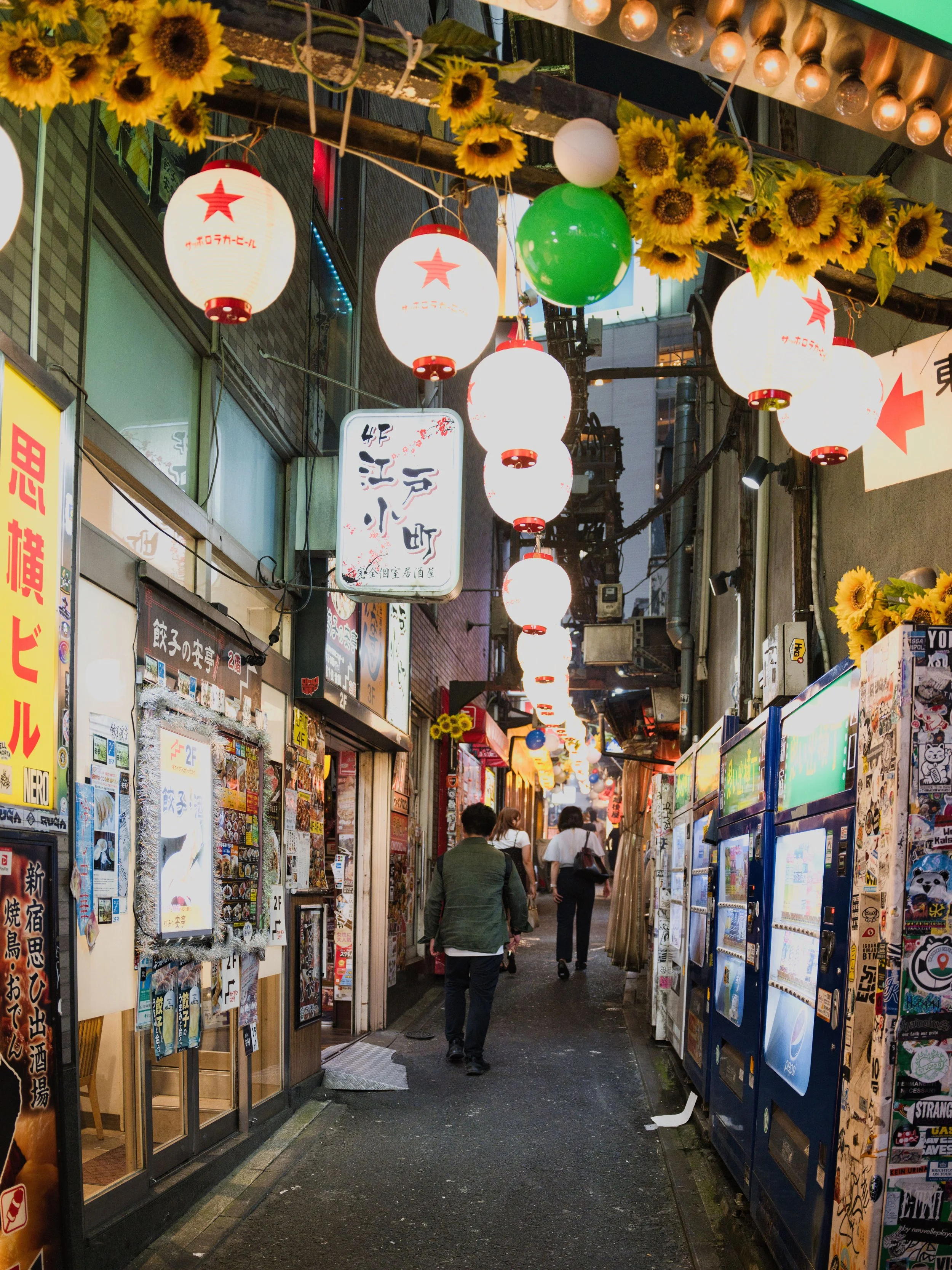 An urban alleyway with Japanese paper lanterns, sunflower decorations, vending machines, and people walking during dusk.