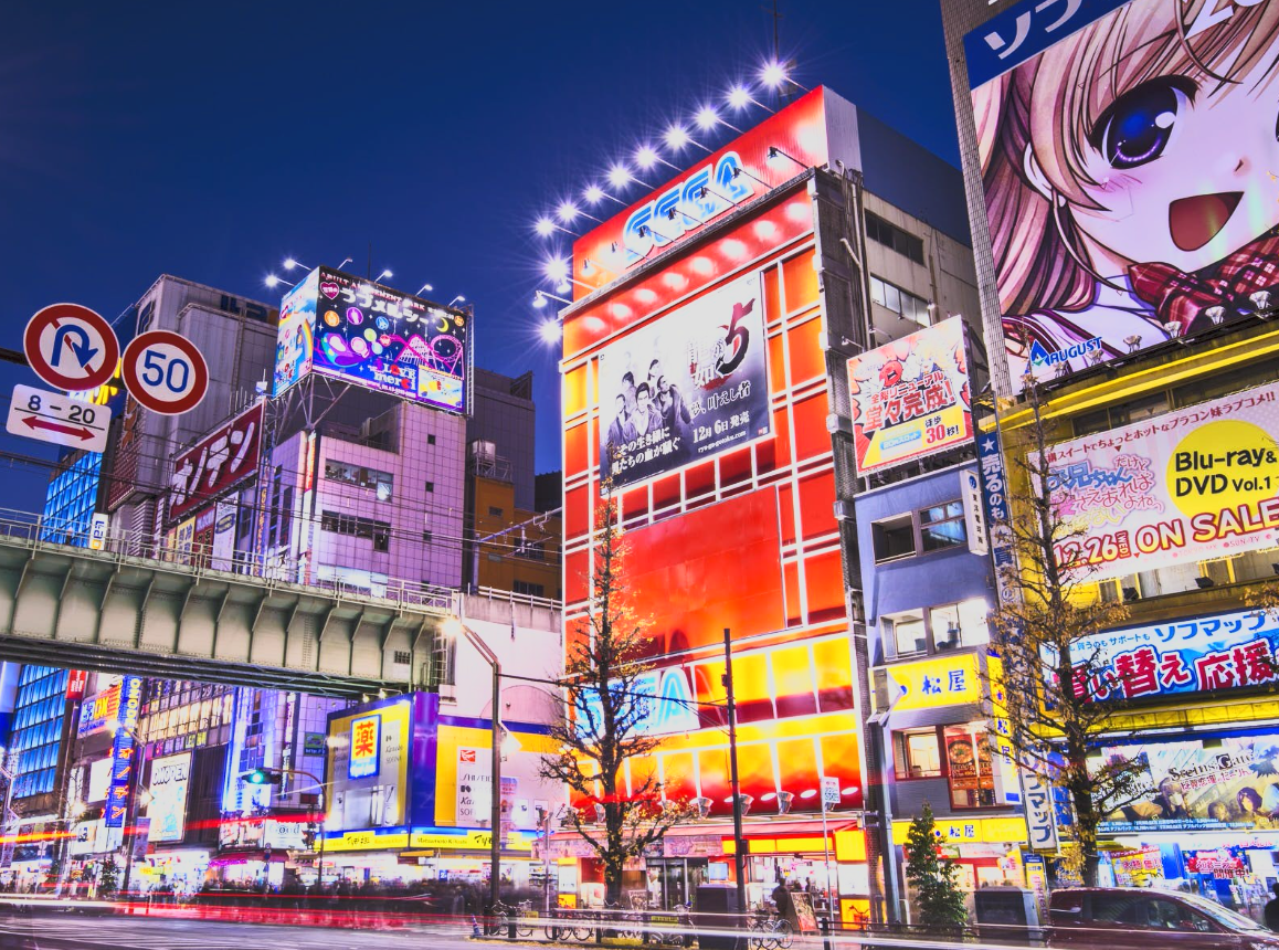 Nighttime cityscape in Japan with brightly lit billboards, signs, and advertisements on tall buildings, featuring colorful graphics and Japanese writing.
