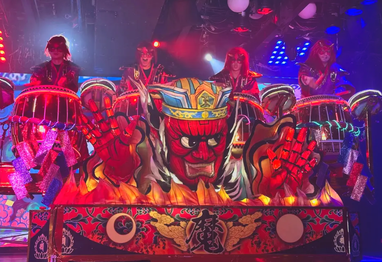 Colorful Japanese-inspired parade float featuring a fierce red face with an angry expression, wearing a blue and yellow hat, surrounded by drums and other ornate decorations, under bright colorful stage lighting.