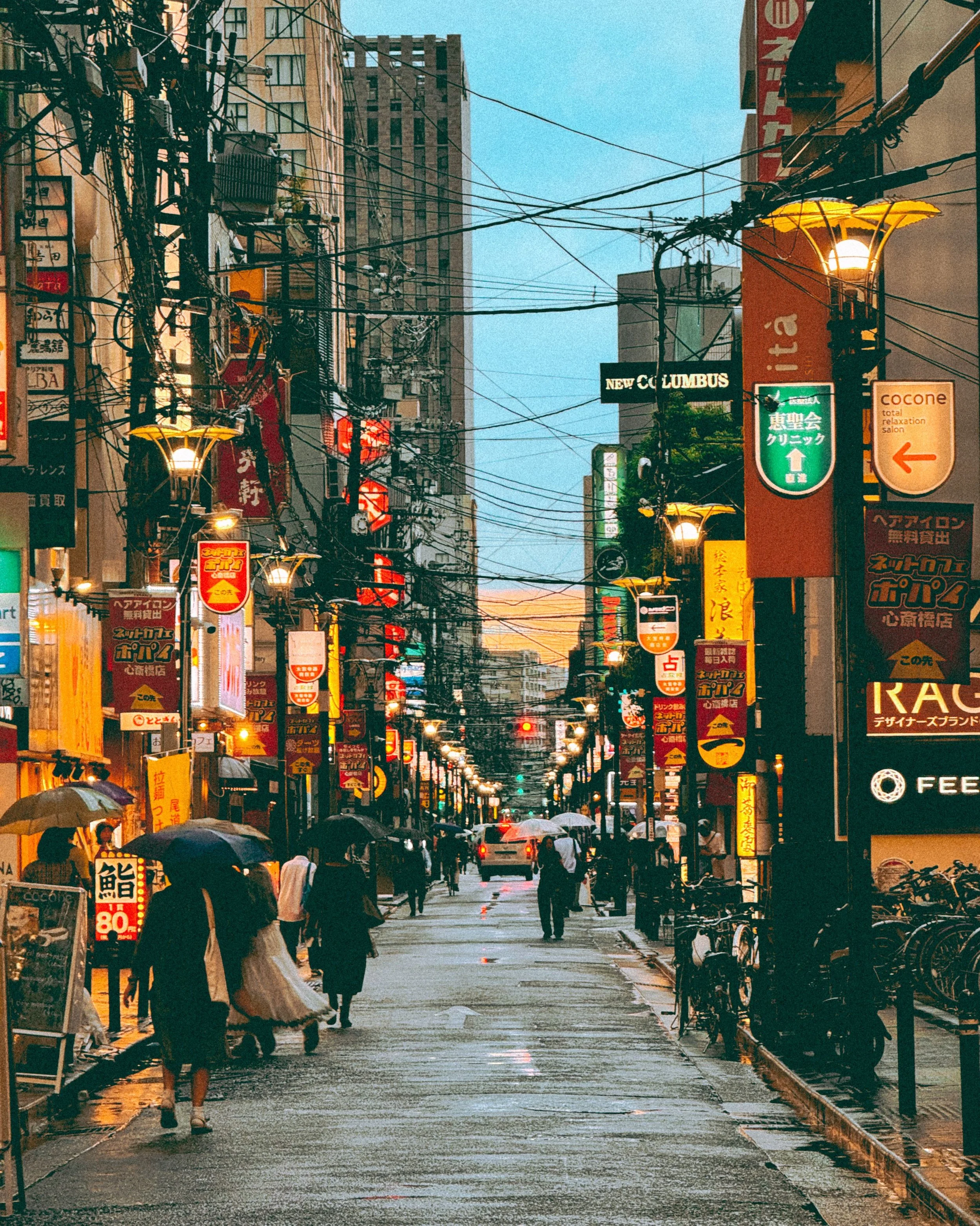 View of a city street at night with illuminated signs in Japanese, including a rounded red lighted sign at the bottom and a taxi driving on the road in the foreground.