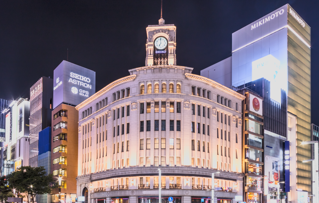 Nighttime city scene featuring a historic building with a clock tower and illuminated modern buildings in the background.