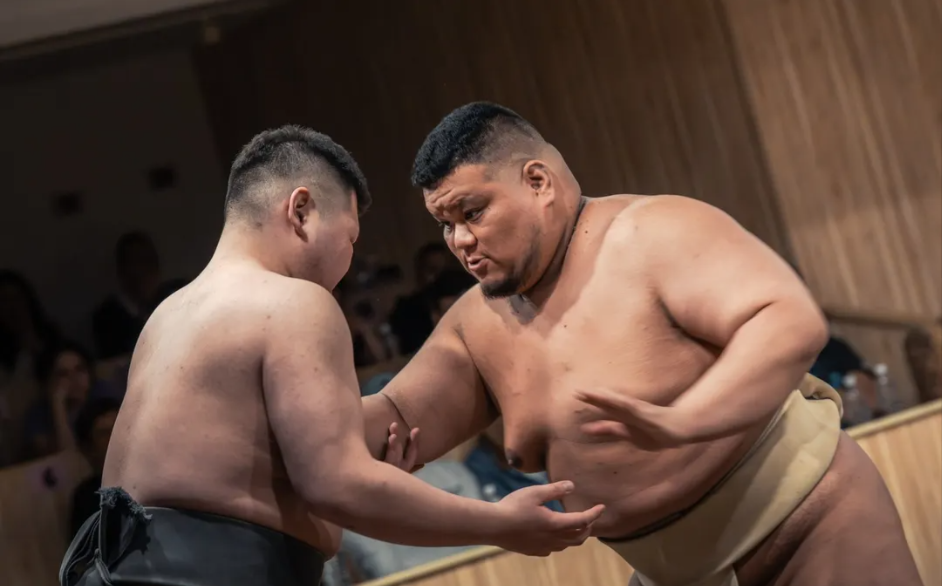 Two sumo wrestlers in a ring, engaging in a match, with spectators watching in the background.