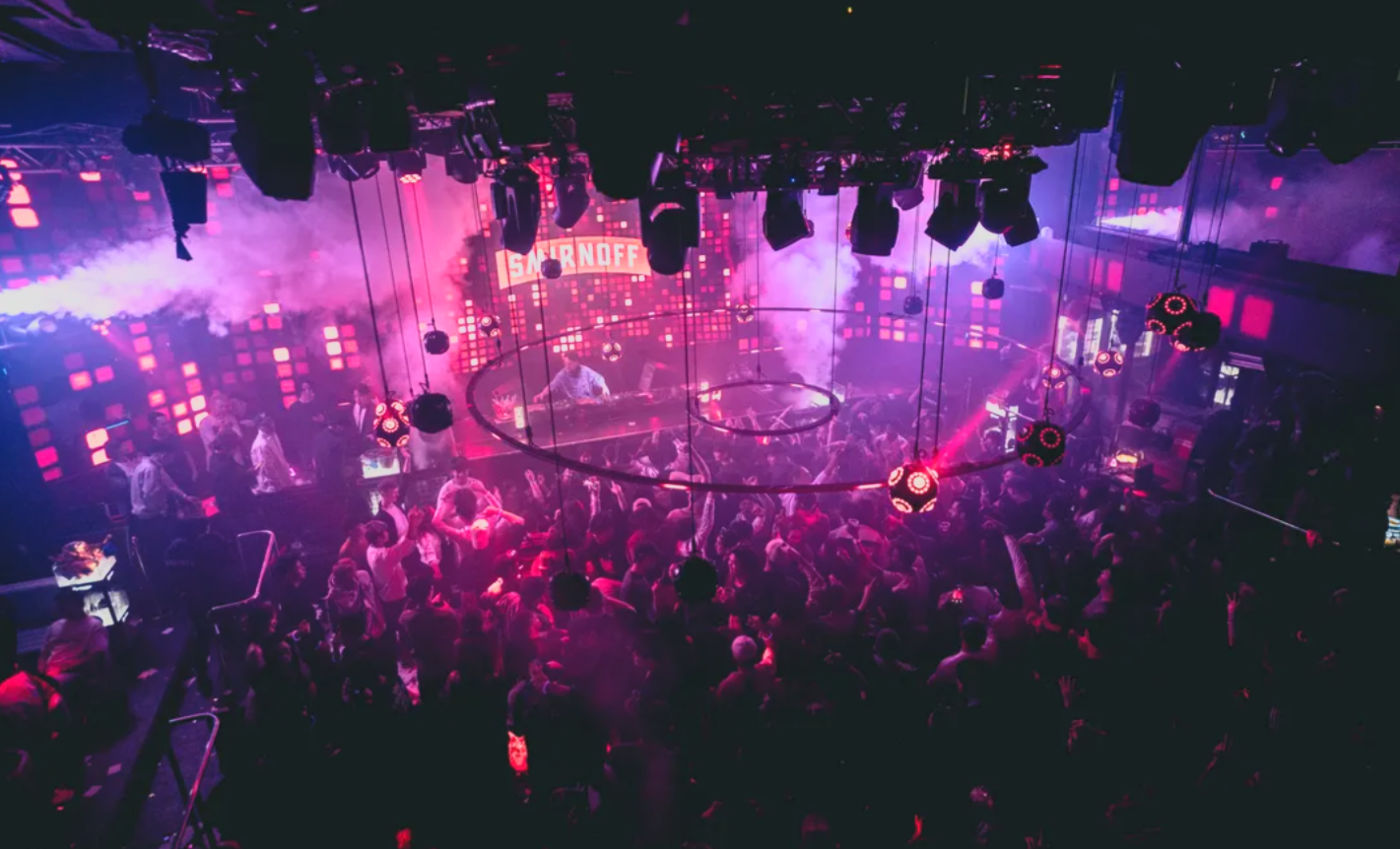 A crowded nightclub dance floor with purple and pink lighting, DJ booth at the back, and a sign that reads 'SEARNOFF' above the DJ. The crowd is dancing under hanging spherical lights.