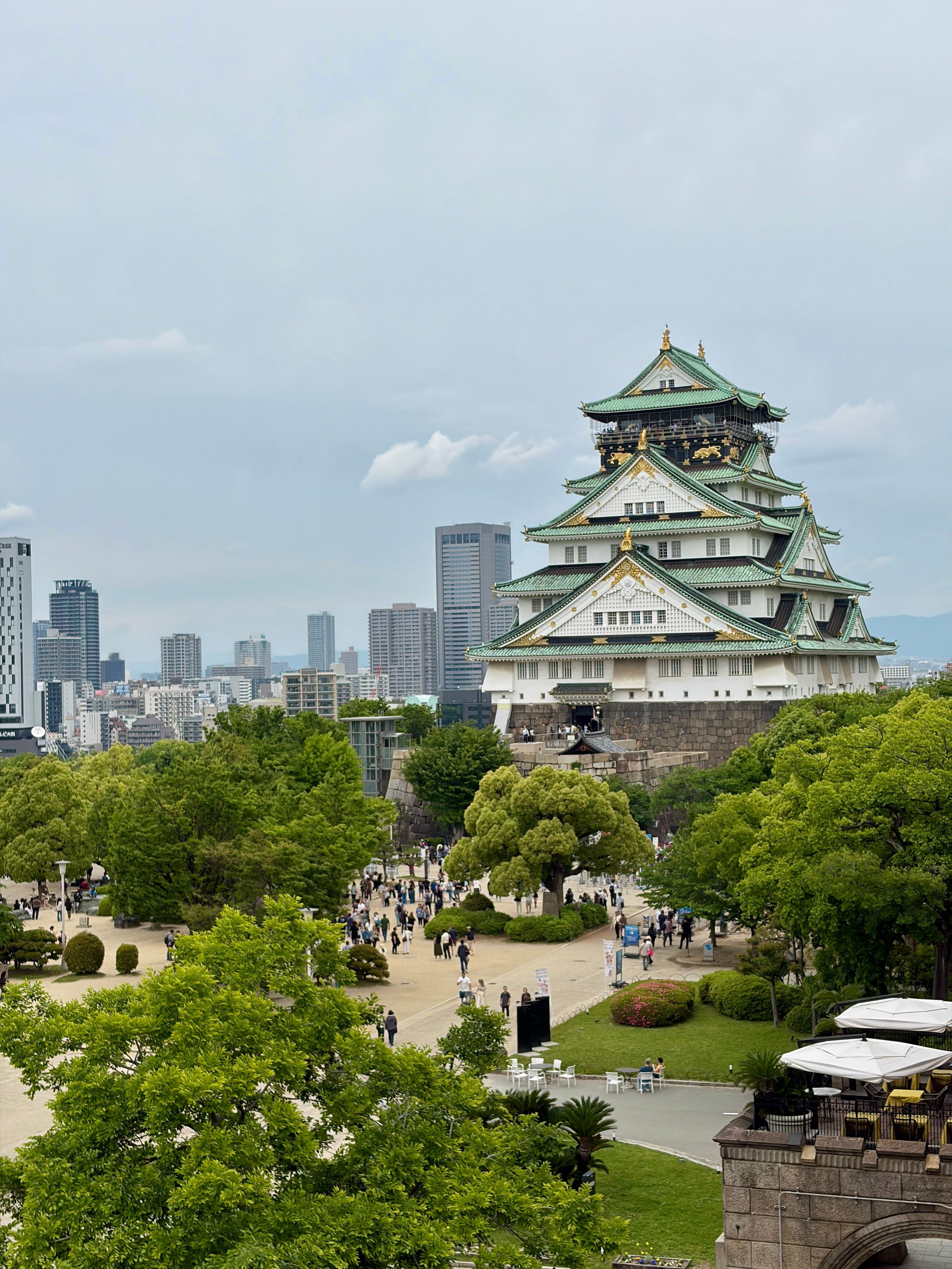 The image features Nagoya Castle with its traditional Japanese architecture, surrounded by lush green trees. In the backdrop, there are modern high-rise buildings and a cloudy sky. Visitors are seen walking and enjoying the park area around the castle.