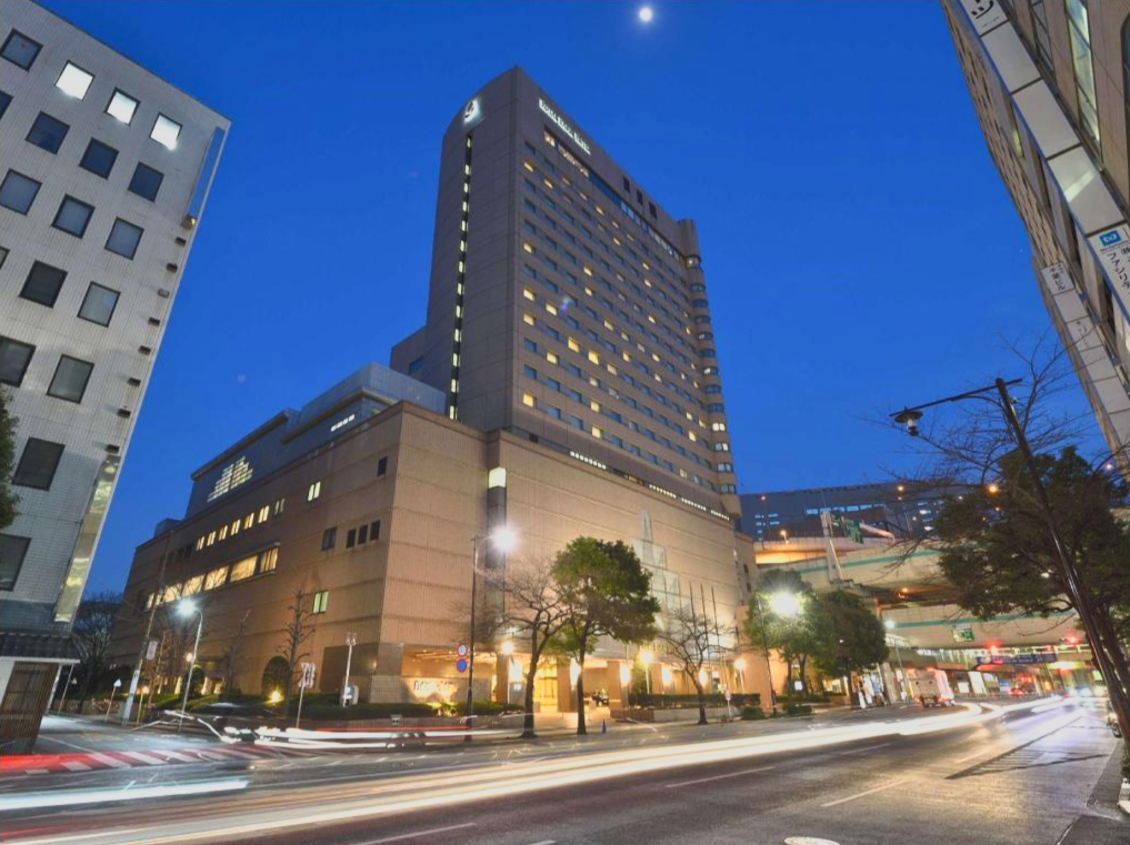 Tall commercial building illuminated at dusk with city streets and light trails from cars in the foreground.