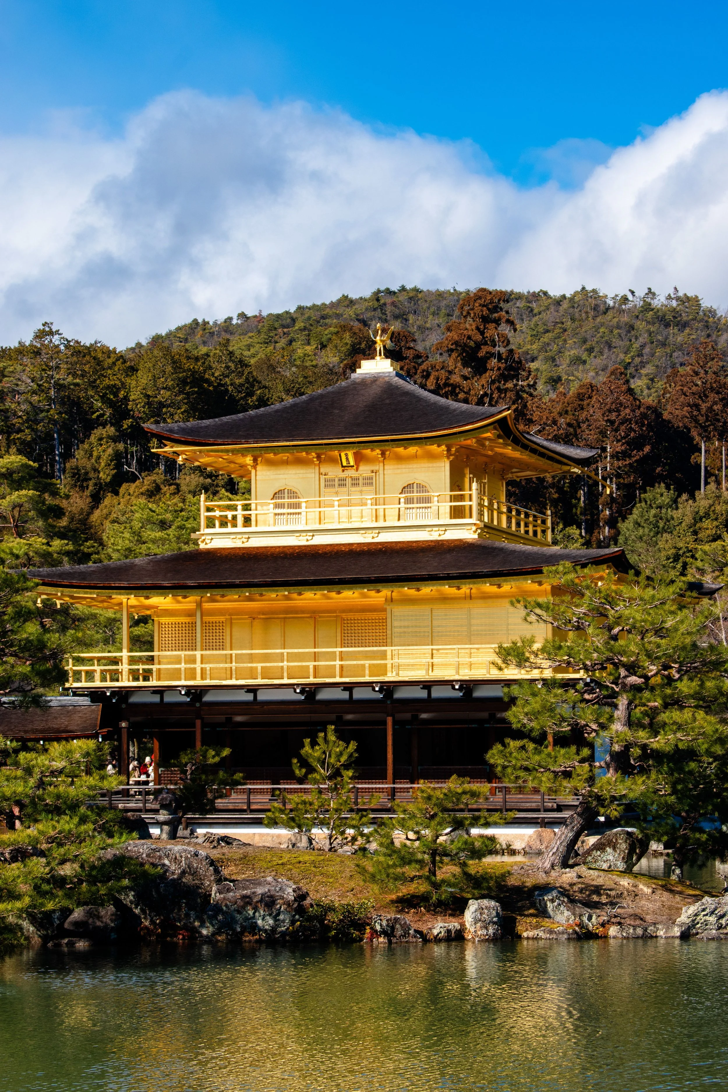 Golden Buddhist temple with traditional architecture, surrounded by trees and rocks, reflecting in a nearby water body, with cloudy sky and mountain in the background.