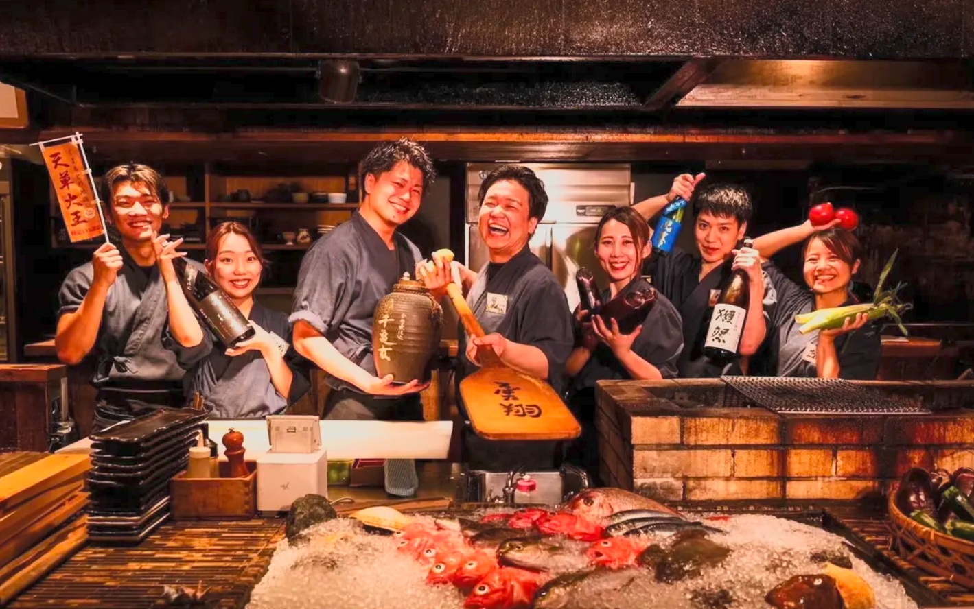 Group of seven in traditional Japanese chef uniforms smiling behind a seafood display with fish on ice, holding various bottles and vegetables, in a restaurant kitchen.