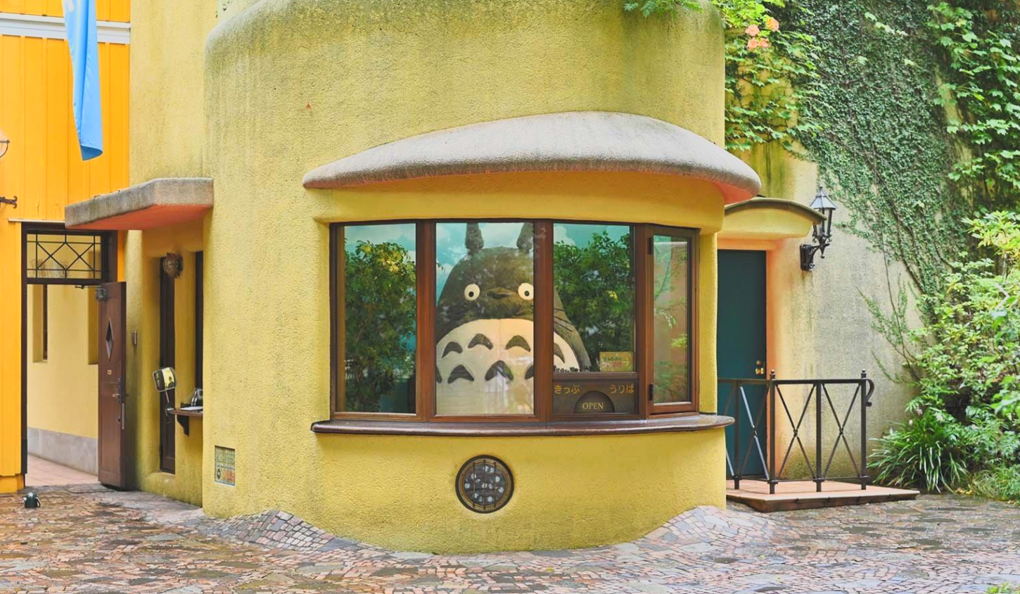 A yellow, rounded building with large window showcasing Totoro character from studio Ghibli. Totoro's face is visible inside, looking out. The setting is garden-like with trees and ivy on the wall.