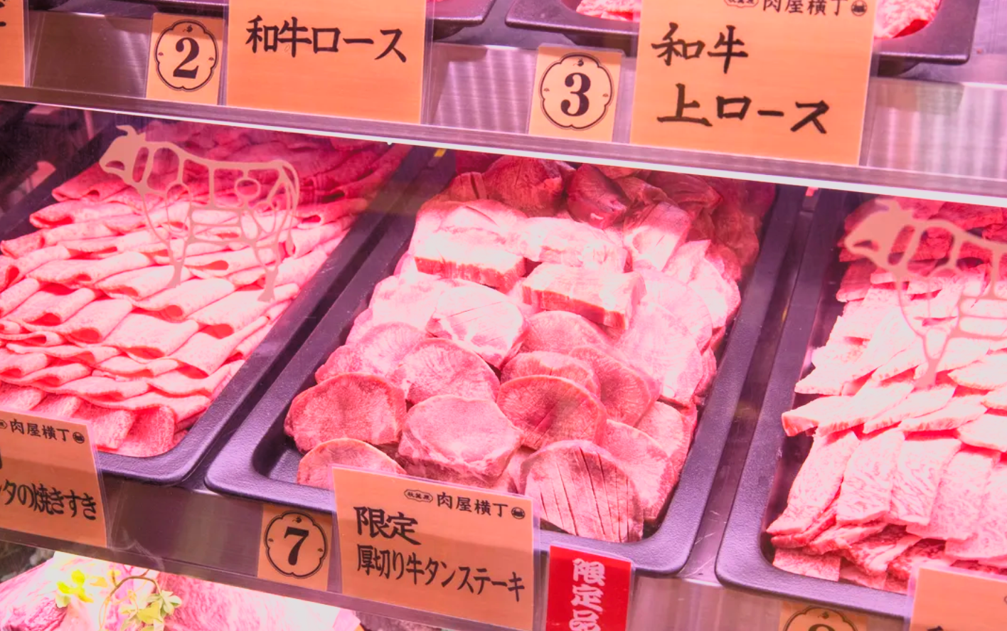Display case with trays of raw meat, including beef steaks and other cuts, with labels in Japanese indicating different types of meat, such as beef loin and sirloin steaks.