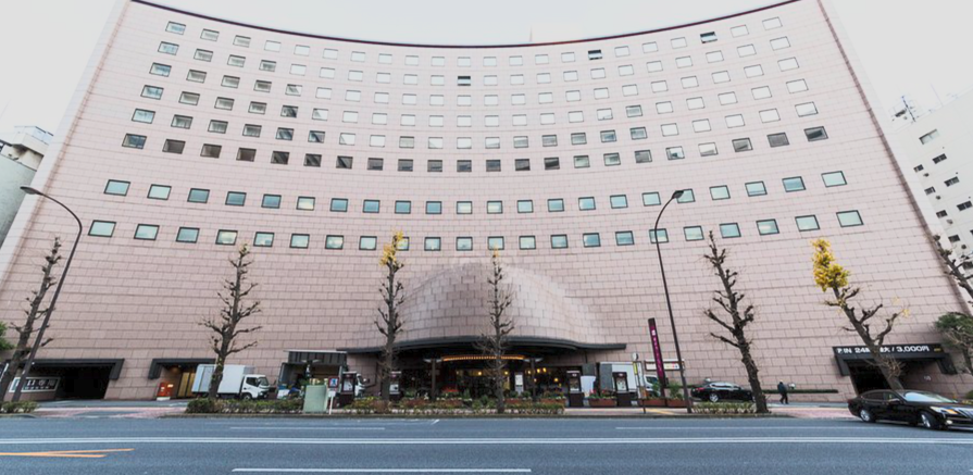 A large curved hotel building with multiple windows, street trees, and parked cars in front.
