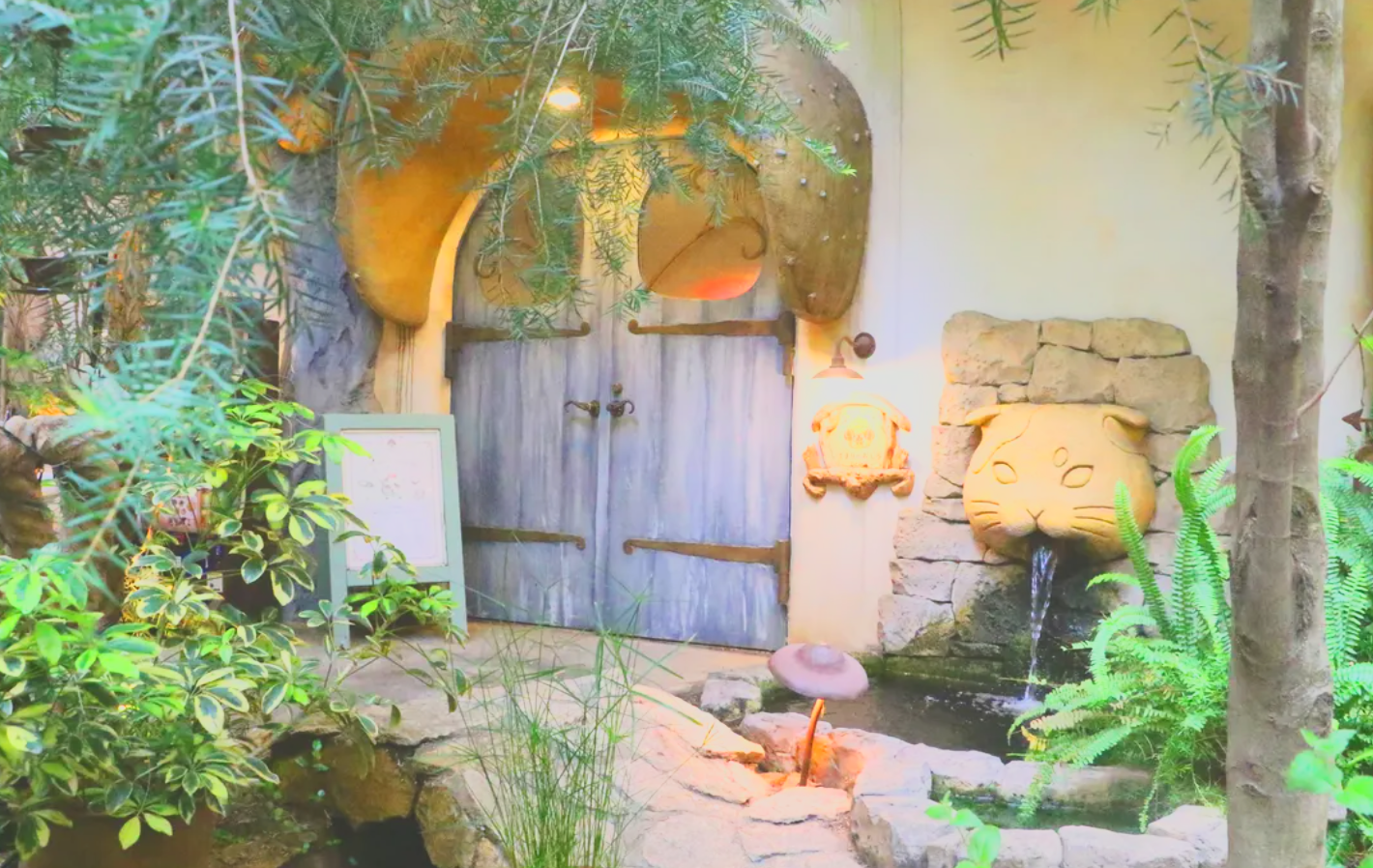 Decorative indoor garden area with a stone wall, a large cat face water fountain, greenery, a small pond, and a bluish wooden door.