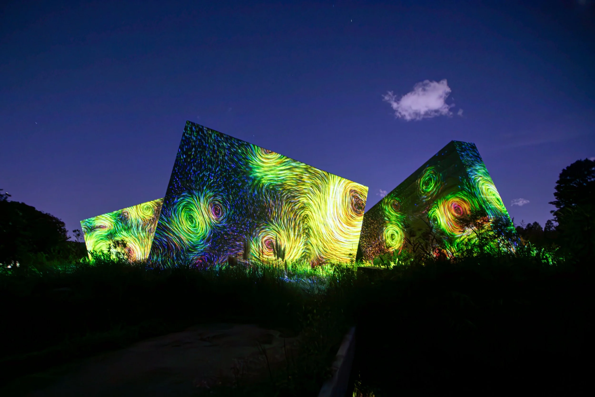 Nighttime outdoor scene with large geometric structures illuminated with colorful swirling light projections, against a dark sky with a few clouds and visible stars.