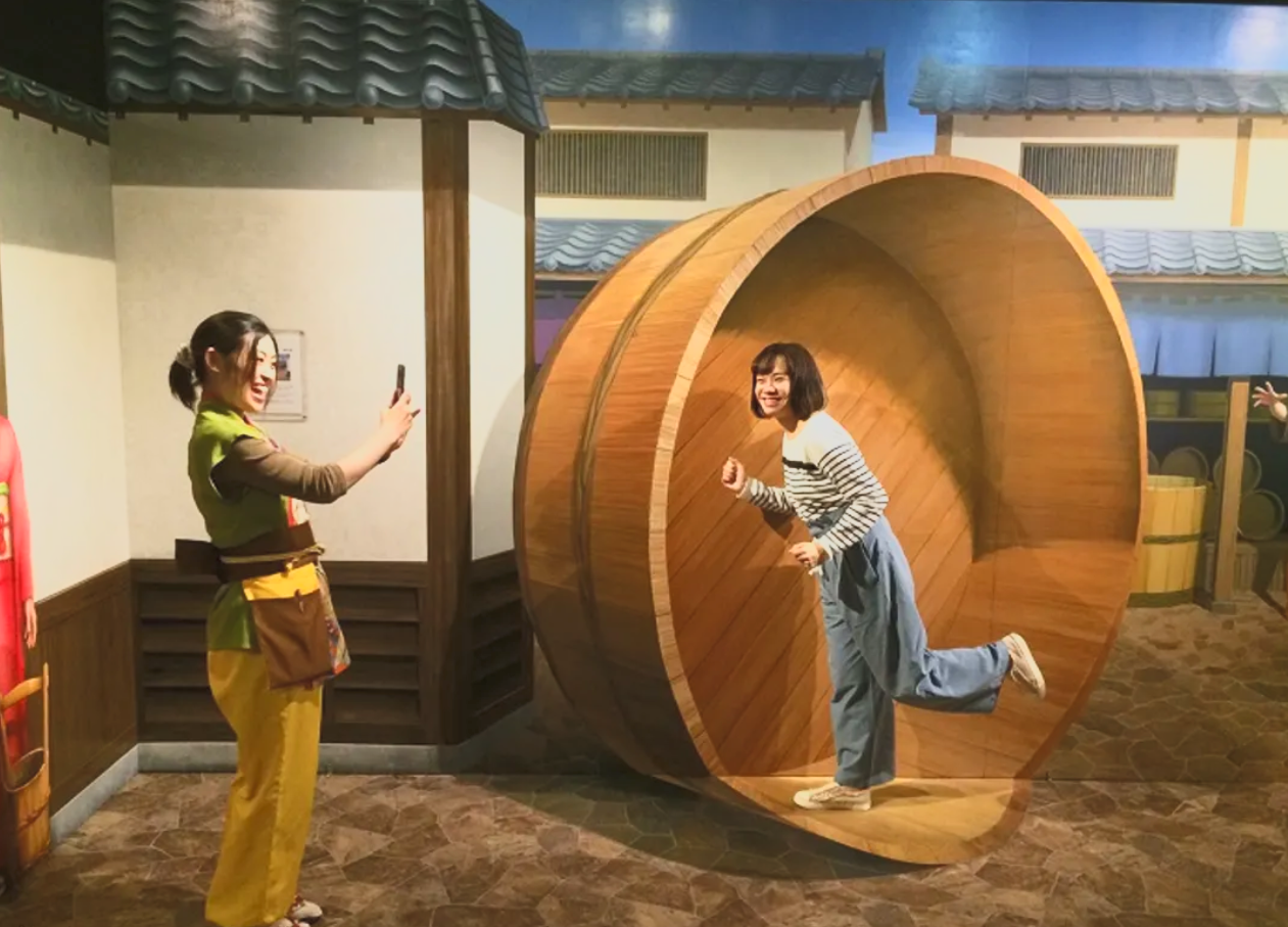 Two women smiling, one taking a photo with her phone while the other leans out of a large wooden circular structure, inside a setting resembling a traditional Japanese building with tiled roofs.